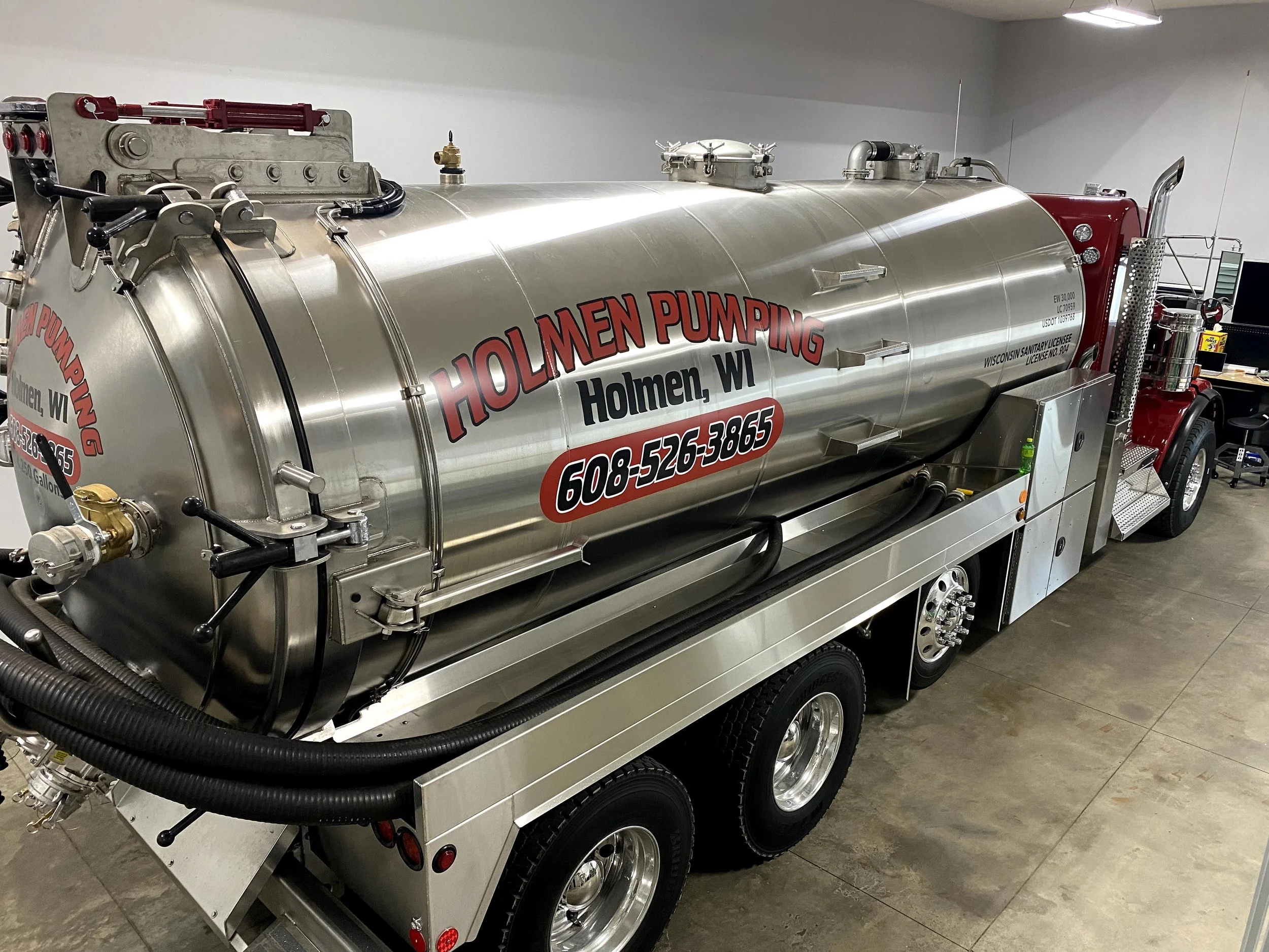 A large stainless steel vacuum truck with company branding for Holmen Pumping, based in Holmen, Wisconsin. The truck has a red cab, four wheels, and various hose attachments, with the company's phone number displayed on the side.