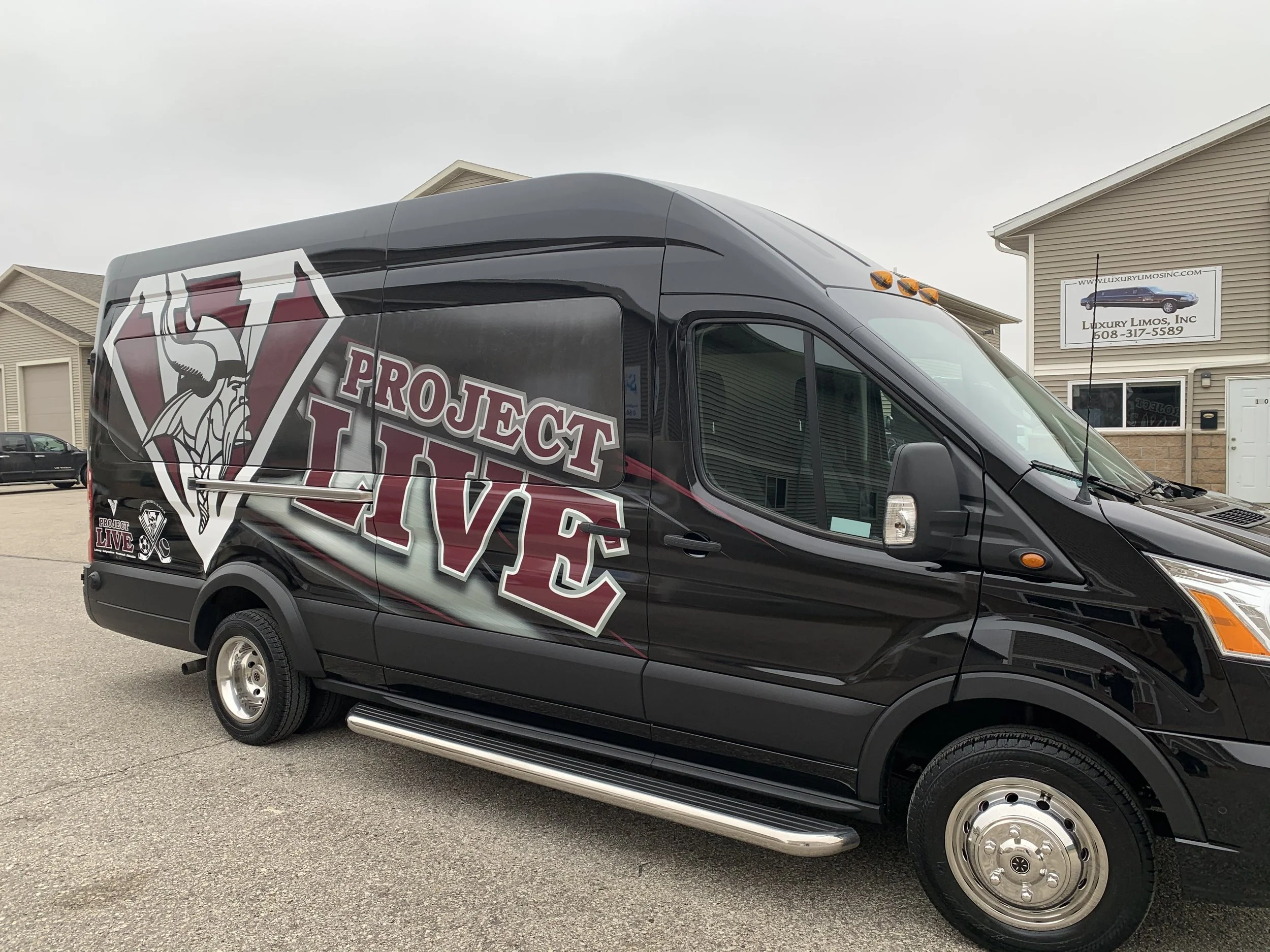 Black van with Project Live logo and a Viking warrior graphic, parked in a lot near beige residential buildings