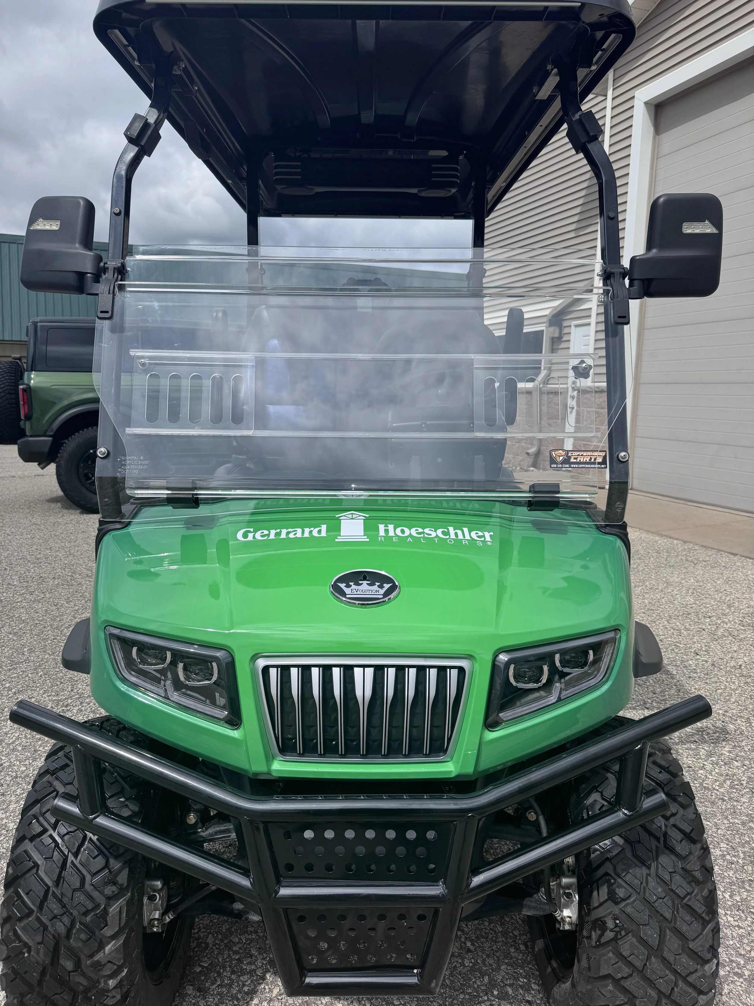 Green golf cart with a clear windshield, black bumper, large off-road tires, and logos for Gerrard Hoeschler Realtors and Evolution on the front.