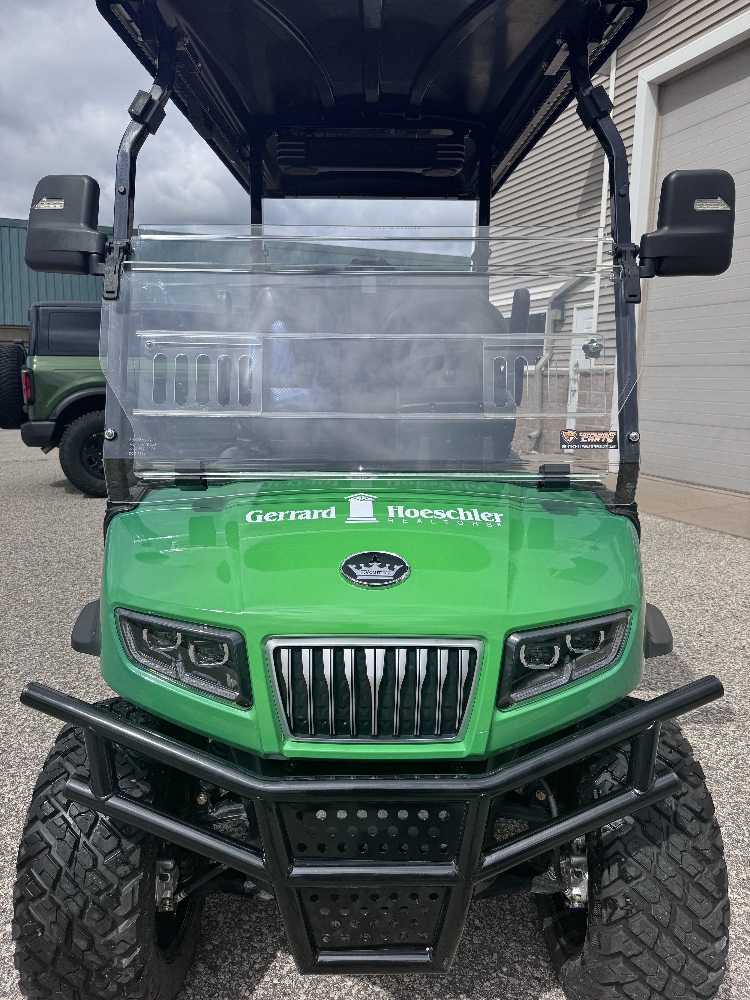 Front view of a green golf cart with a clear windshield, black bumper, and large tires, marked with various sponsor decals, parked in a driveway.