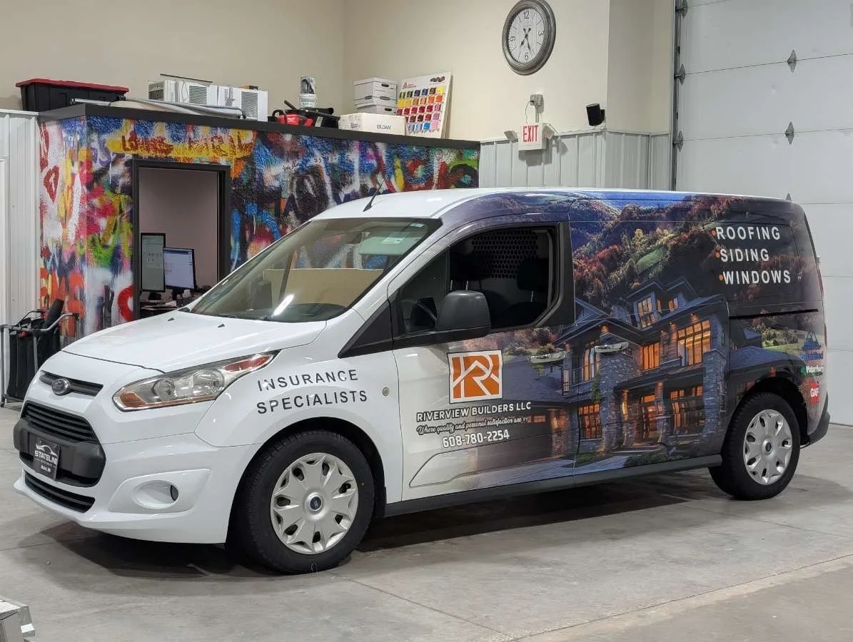 A white commercial van parked inside a garage with a colorful graffiti wall in the background. The van has graphics advertising roofing, siding, and windows services and features the logo and contact information for Riverview Builders LLC.
