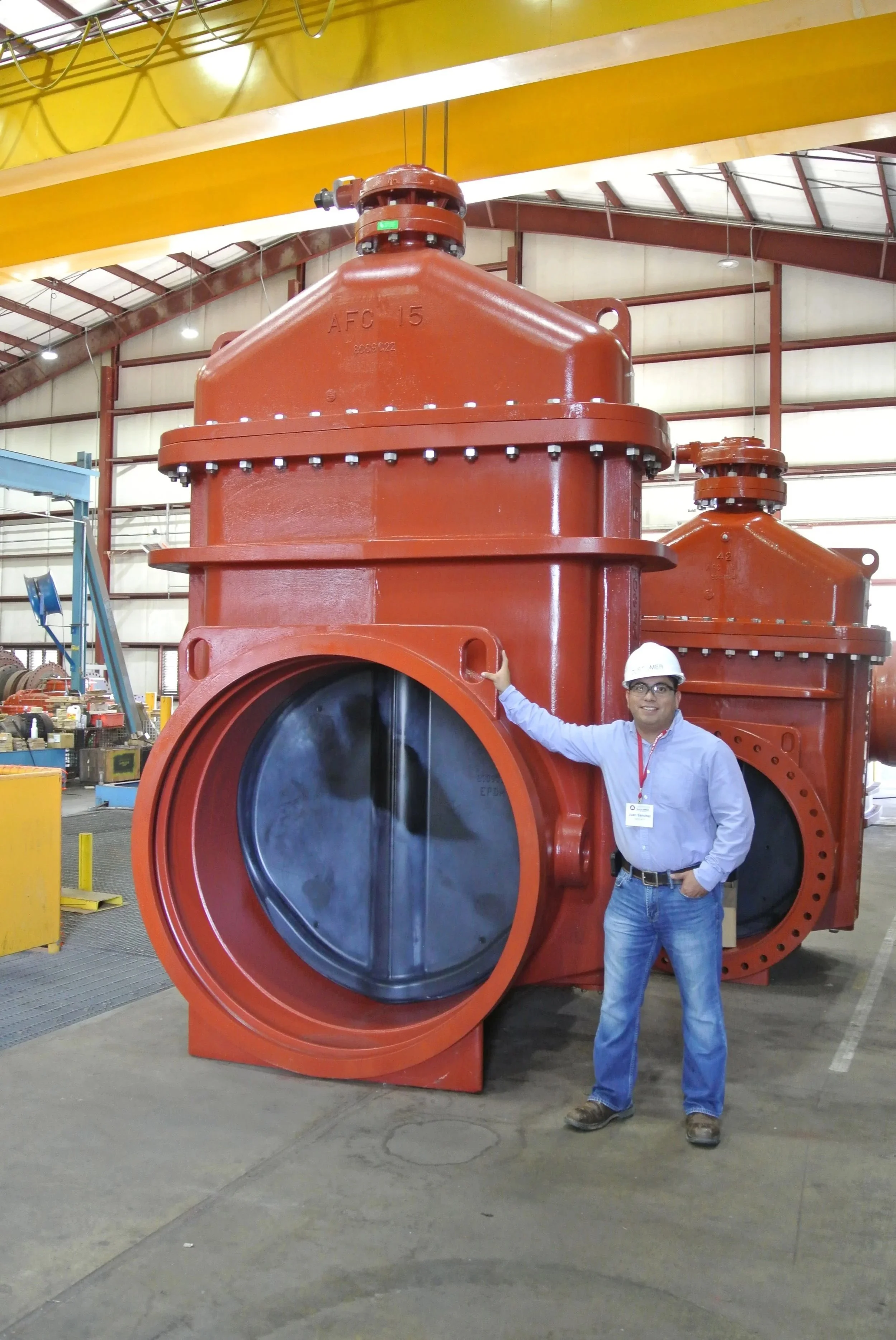 A man wearing safety glasses, a white hard hat, a blue shirt, and jeans, standing next to a large red industrial valve inside a factory or warehouse.
