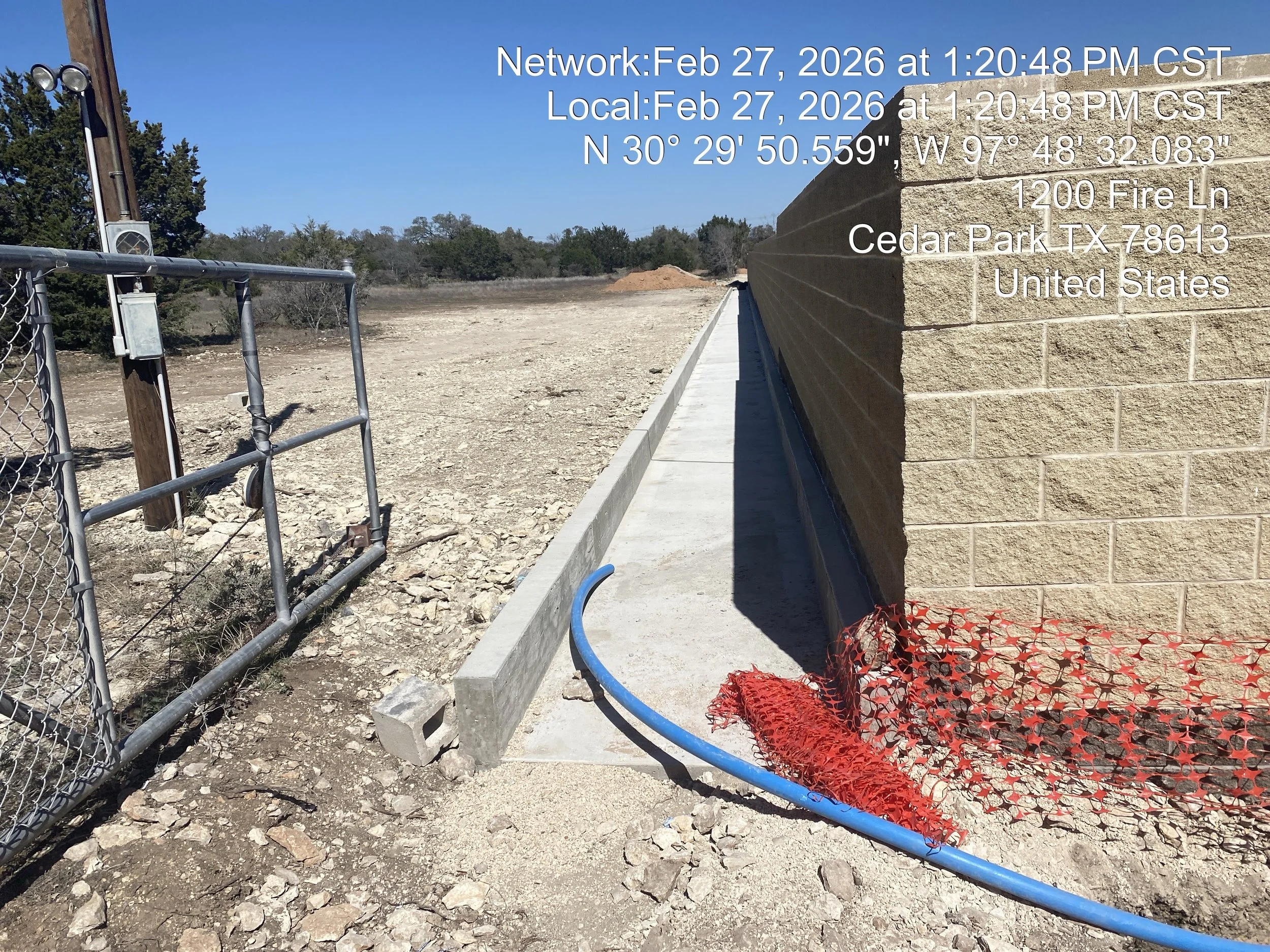 Empty construction site with a concrete sidewalk along a brick wall, a blue hose, orange safety netting, and a chain-link fence, under a clear blue sky.