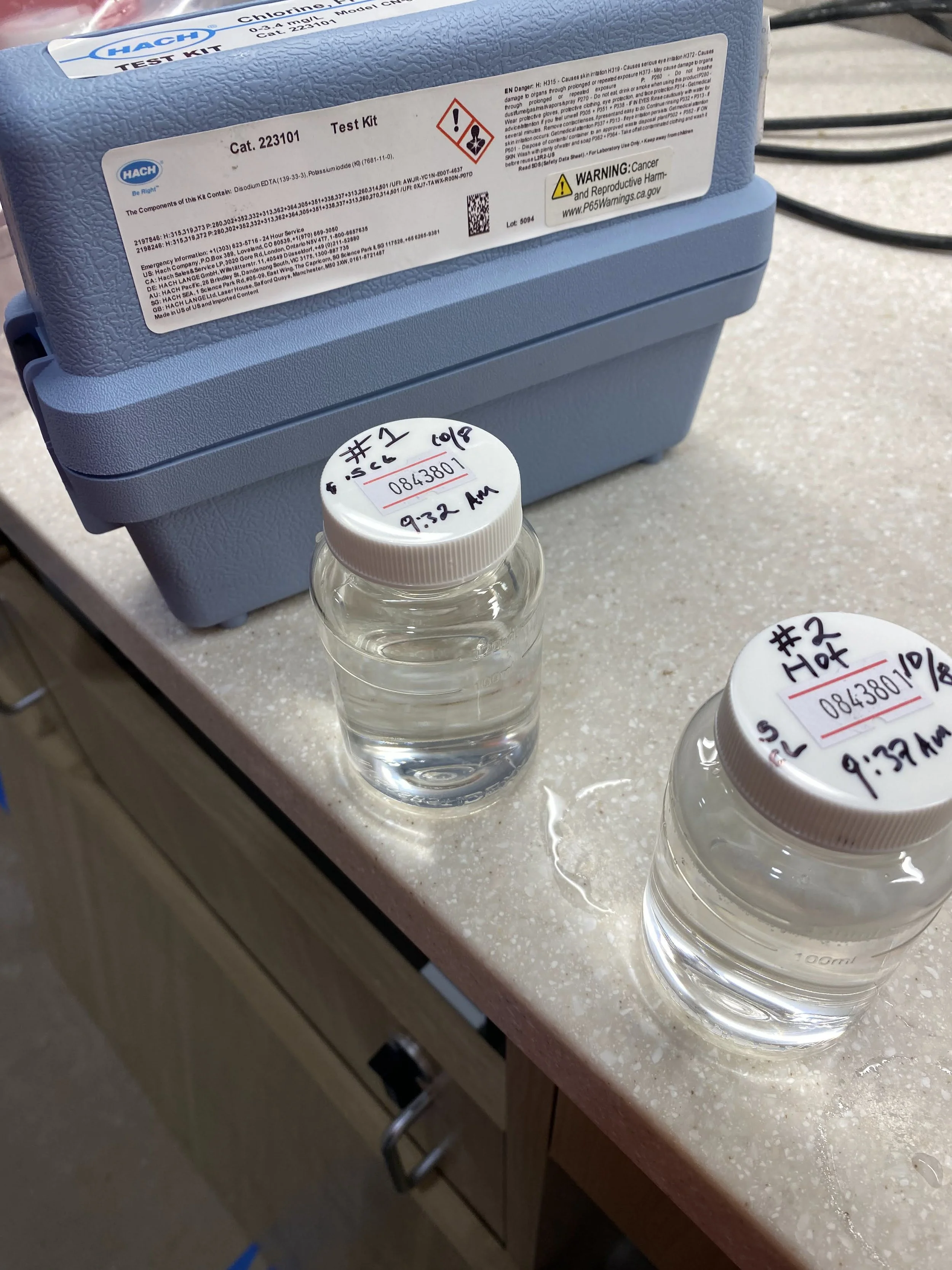 Two small glass jars filled with clear liquid, labeled with handwritten notes and caps, placed on a beige countertop. A blue test kit box with labels and warnings in the background.