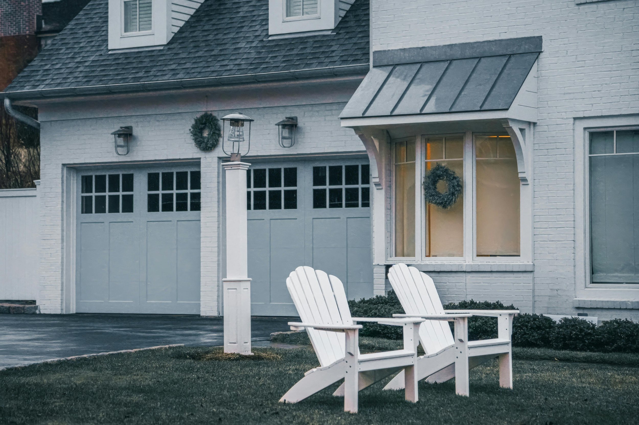 White house with wreaths on the windows and a lantern, two white Adirondack chairs on a grassy lawn in front.