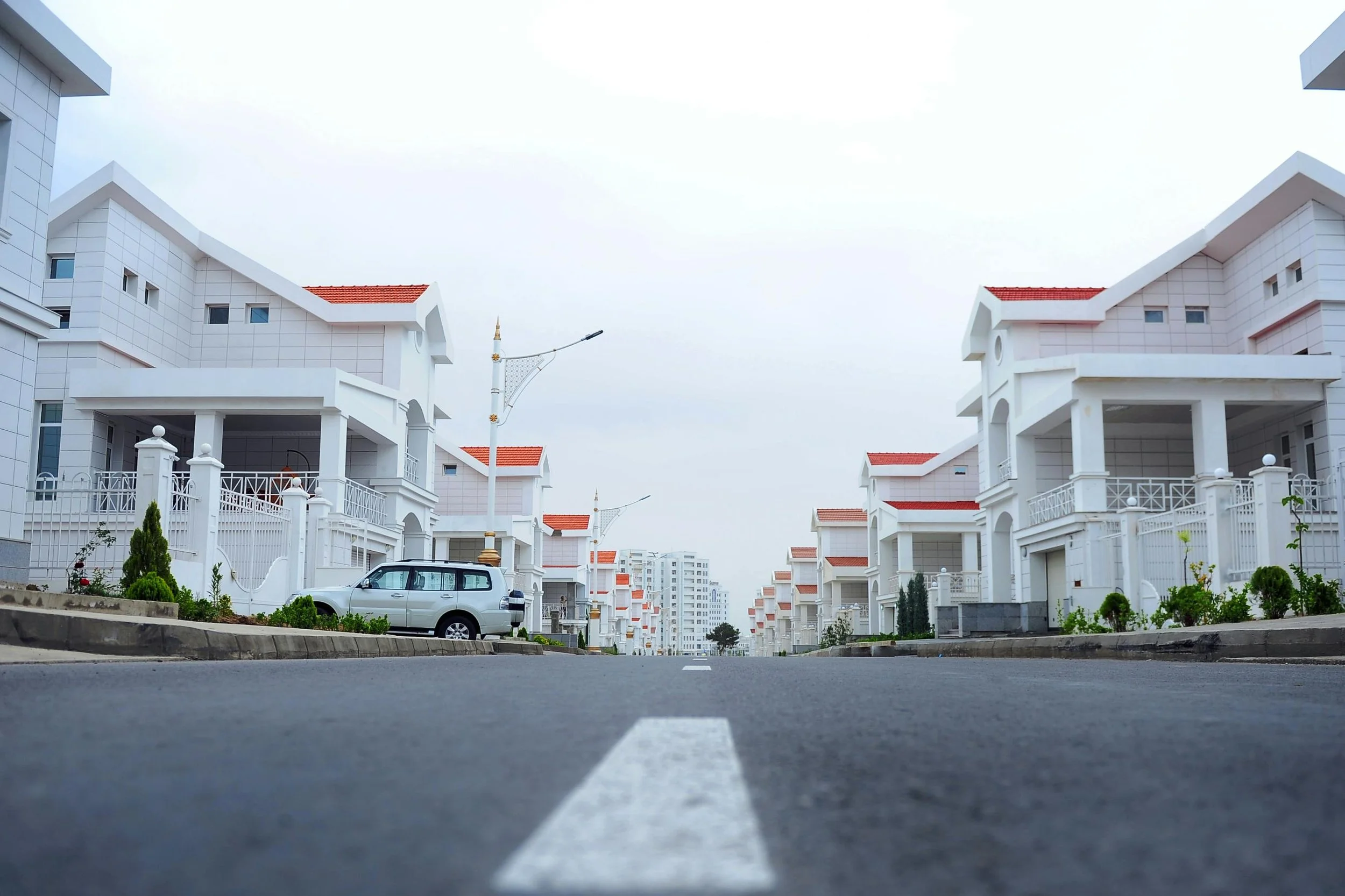 A quiet residential street lined with modern white houses with red-tiled roofs and manicured lawns, viewed from the road level.