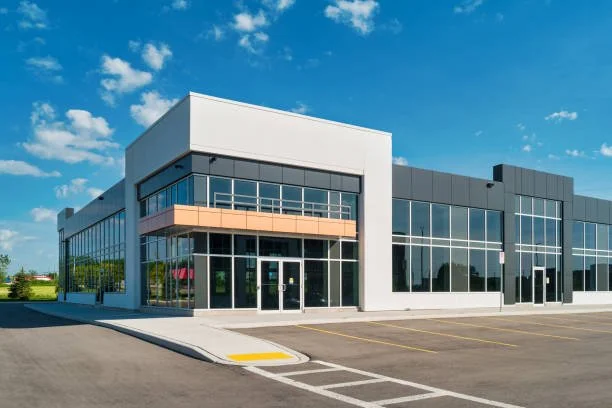 Empty modern commercial building with large glass windows and an adjacent parking lot under a partly cloudy sky.