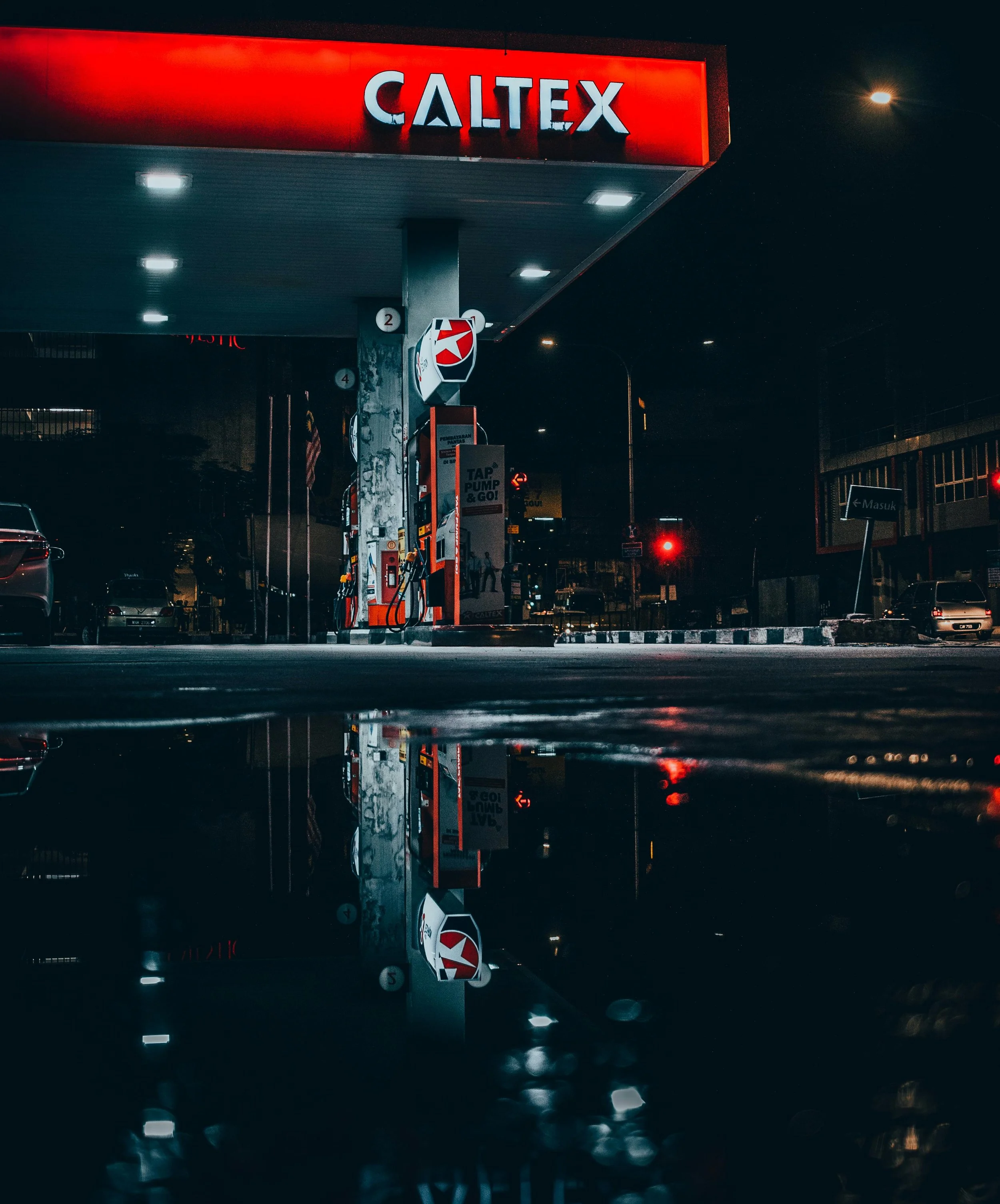 Night scene of a Caltex gas station with illuminated signage, gas pumps, and cars parked around, reflected in a puddle on the ground.