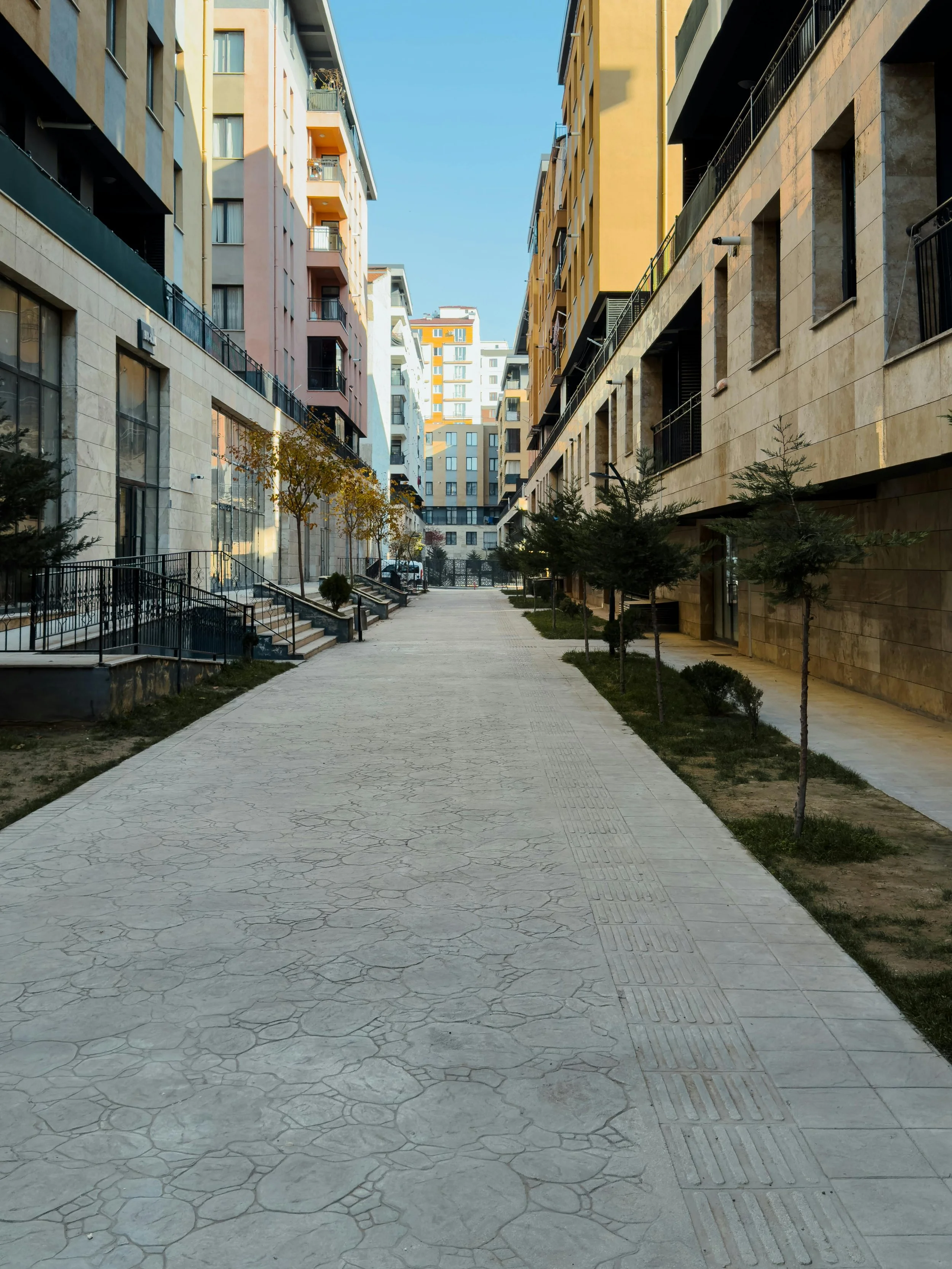 A modern city street with tall residential buildings on both sides, a paved walkway, small trees, and some vehicles in the distance under a clear blue sky.
