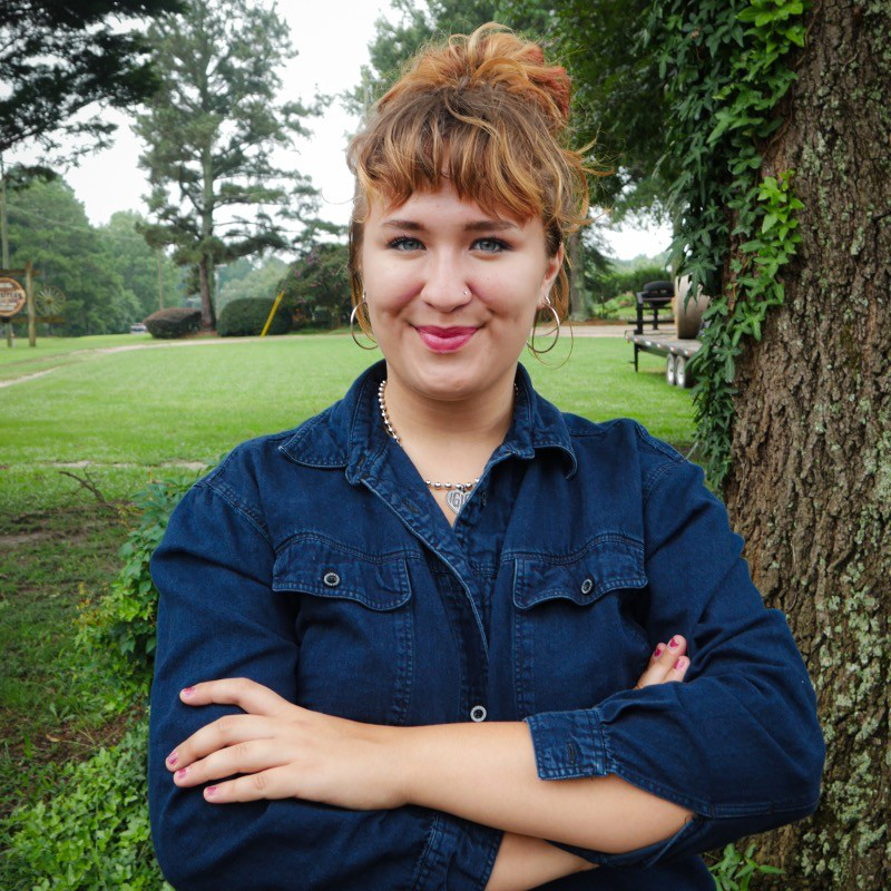 A young woman with short, curly, reddish-brown hair and light skin, standing outdoors with her arms crossed, smiling at the camera, next to a large tree with green foliage, in a park setting.