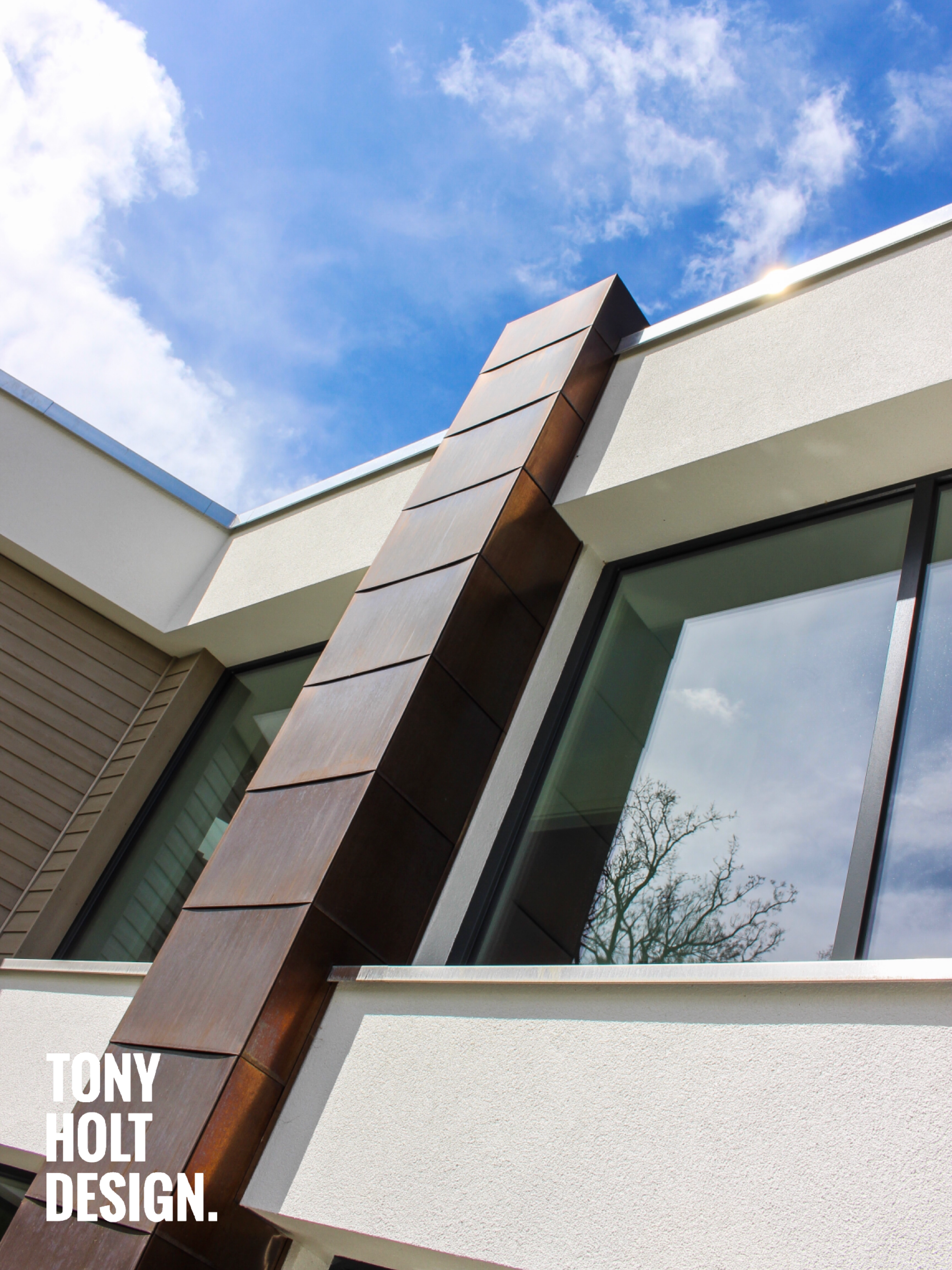 Close-up of a modern house exterior featuring a copper-clad feature column, white render walls, composite cladding, and floor-to-ceiling aluminium windows, designed by Tony Holt Design, a CIAT-registered architectural practice.