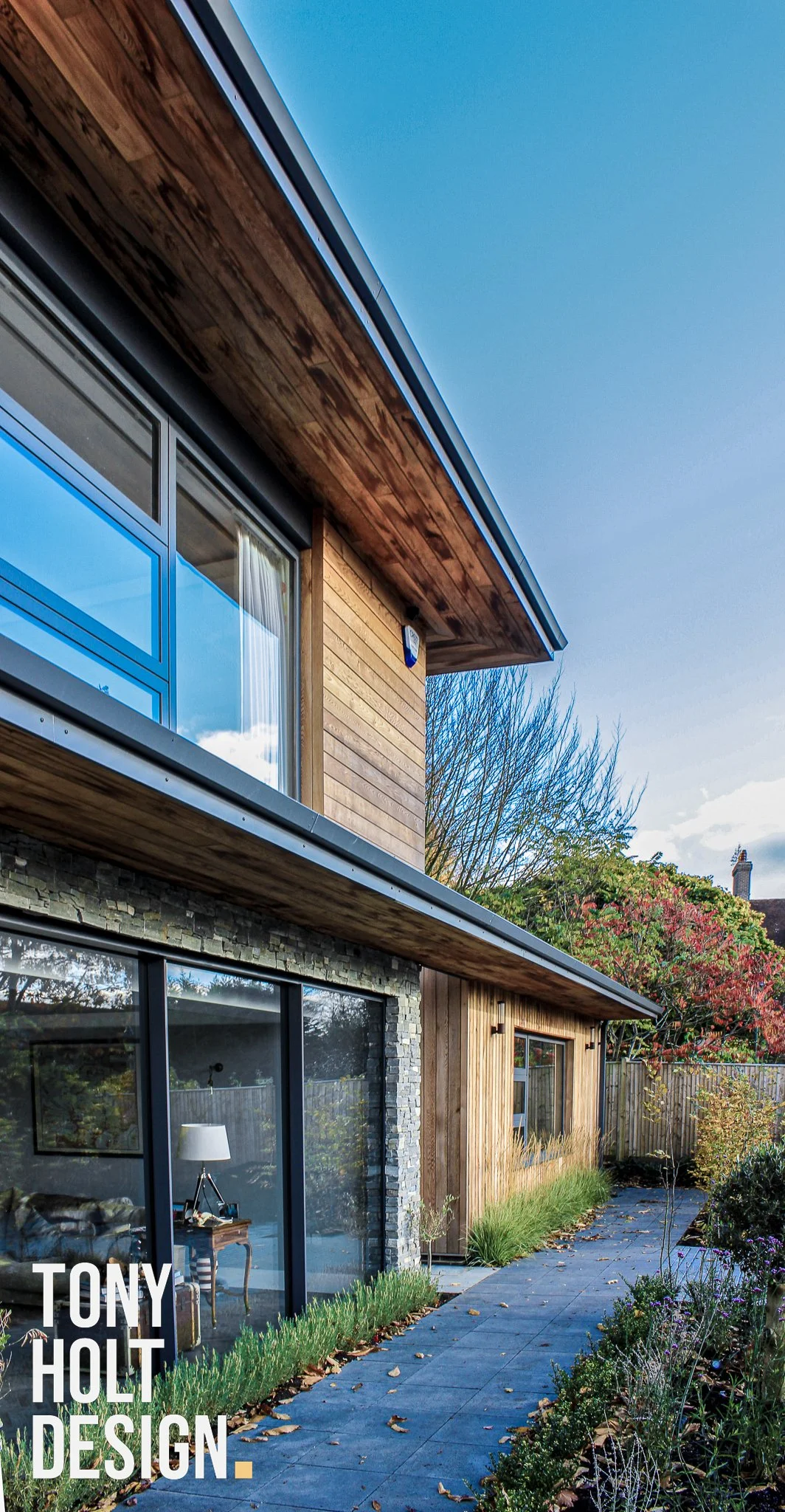 Modern two-story house with large glass sliding doors, wooden and stone exterior, and a landscaped garden with walkway, trees, and shrubs, designed by Tony Holt Design, a CIAT-registered architectural practice.