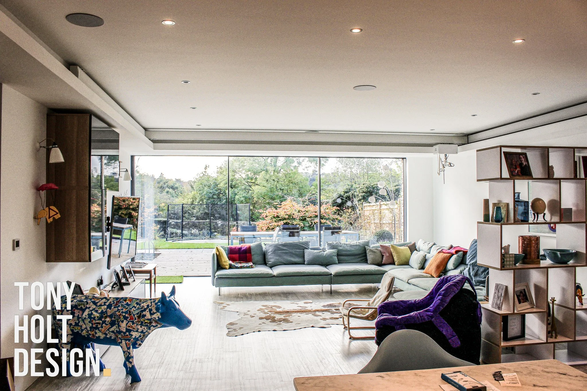 Spacious modern living room featuring a light-colored sectional sofa with colorful cushions, cow-shaped sculpture, and white-and-wood open shelving. Large glass sliding doors open onto an outdoor patio, designed by Tony Holt Design, a CIAT-registered