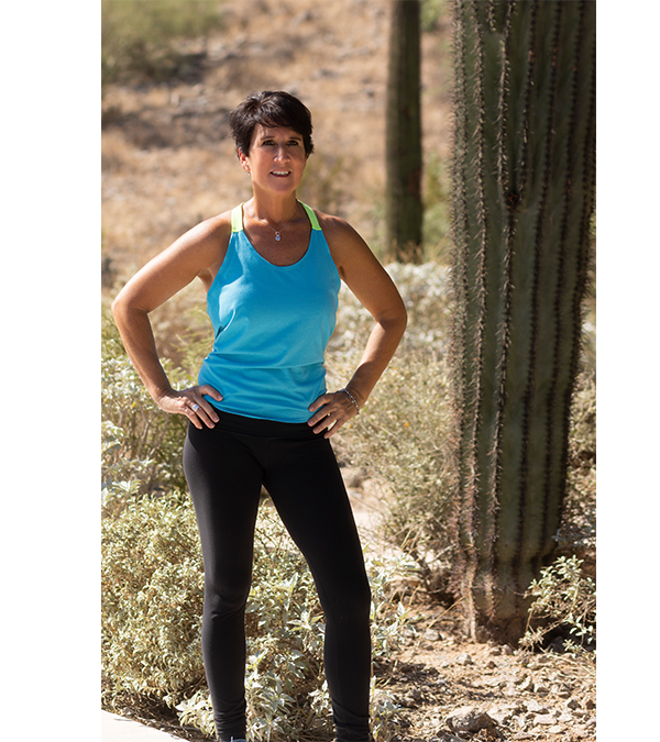A woman with short dark hair standing outdoors in a desert landscape with cactus plants, wearing a blue athletic tank top and black leggings, smiling with hands on her hips.