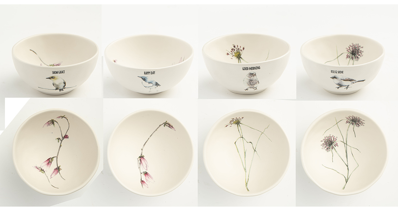 Eight white ceramic bowls with botanical illustrations of flowers and birds, with phrases like "Show Grace," "Happy Day," "Good Morning," and "Rise & Shine" labeled on some bowls. Some bowls are empty, while others display delicate floral designs.