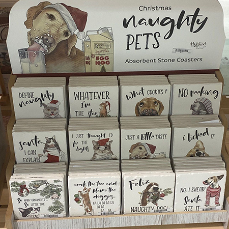 Display of Christmas-themed naughty pet coaster sets with humorous phrases and animal illustrations, arranged in rows on a shelf.