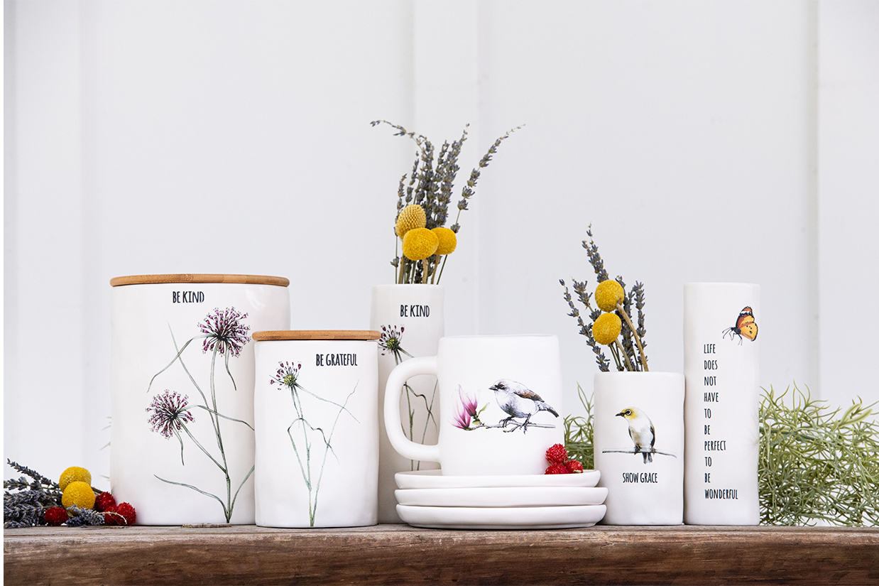 A collection of decorated white ceramic mugs and containers with floral and bird designs, accompanied by sprigs of lavender, yellow billy balls, and dried greenery on a wooden surface.