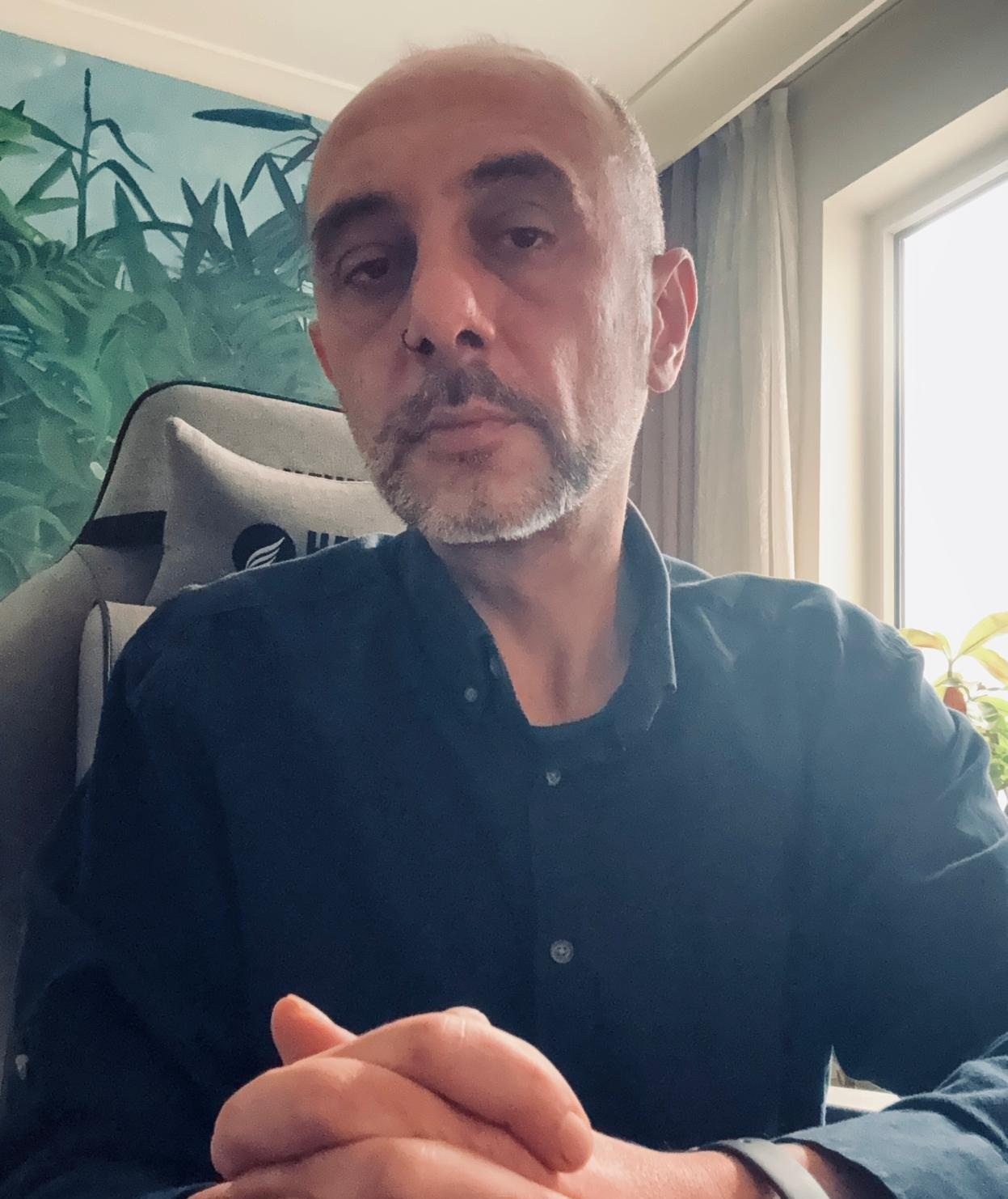 A man with a beard and mustache sitting indoors near a window, wearing a dark button-up shirt, with a bookshelf and green plant in the background.