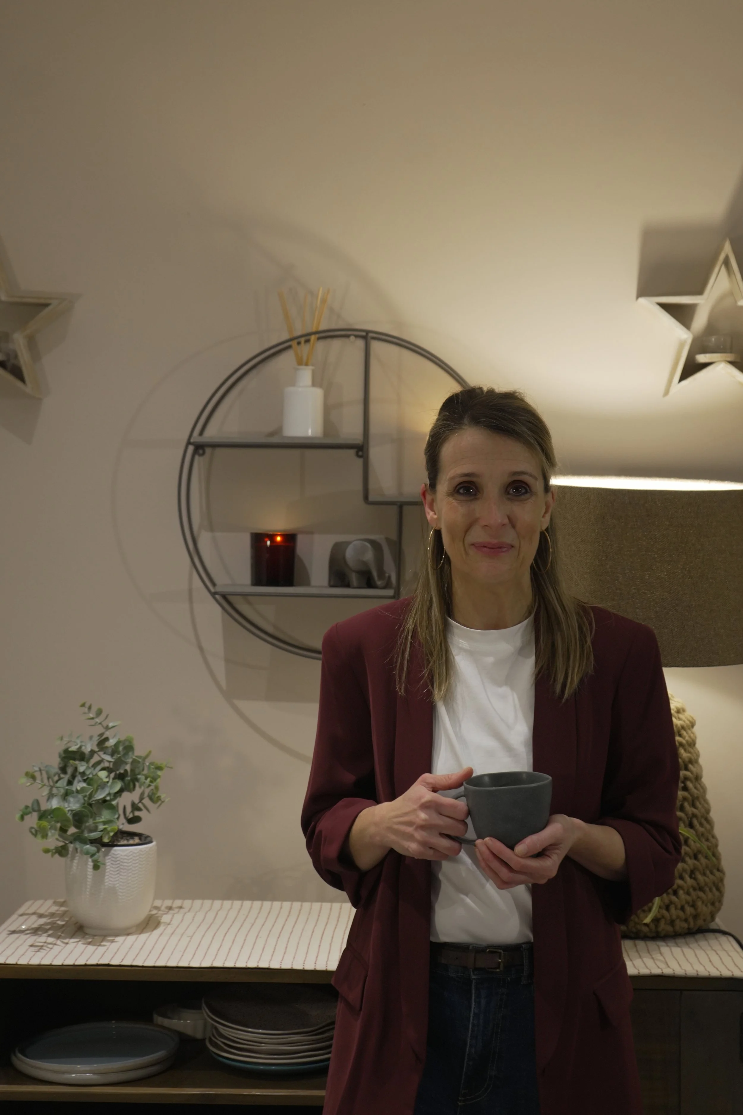 Yvonne (The Intentional Learner) with long brown hair and hoop earrings holding a gray mug, standing in a cozy, decorated room.
