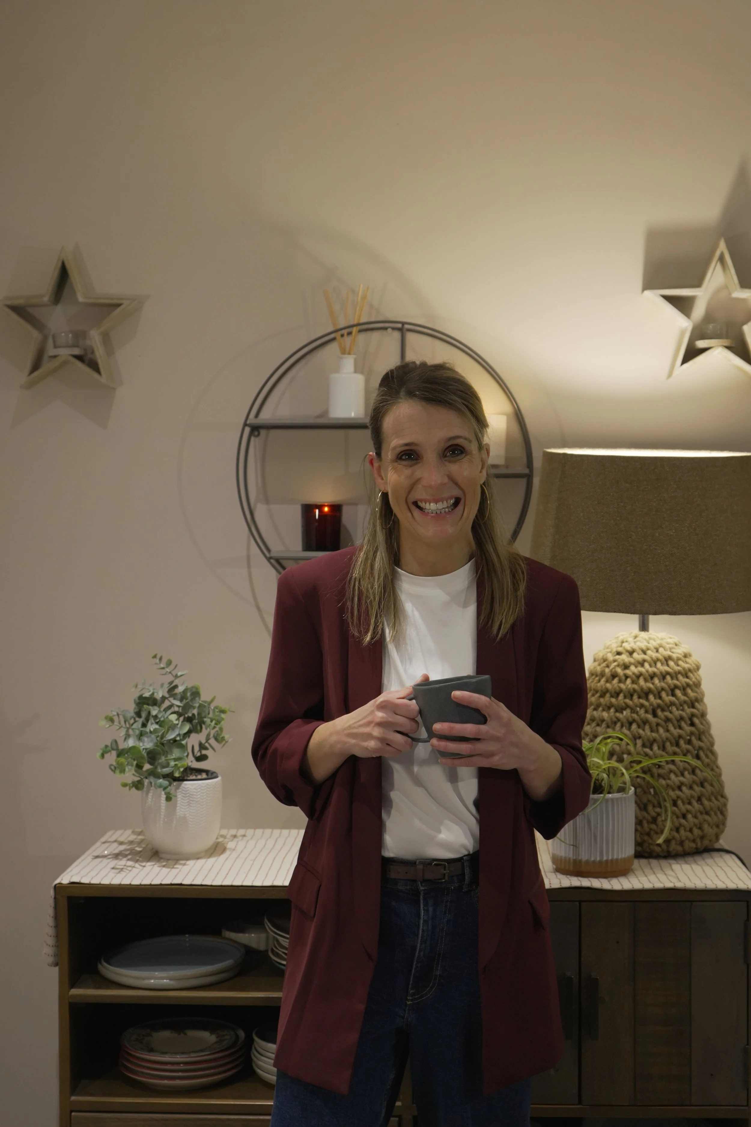 Yvonne (The Intentional Learner) smiling and holding a mug, standing in a cozy room with a sideboard, houseplants, and decorative wall shelves.