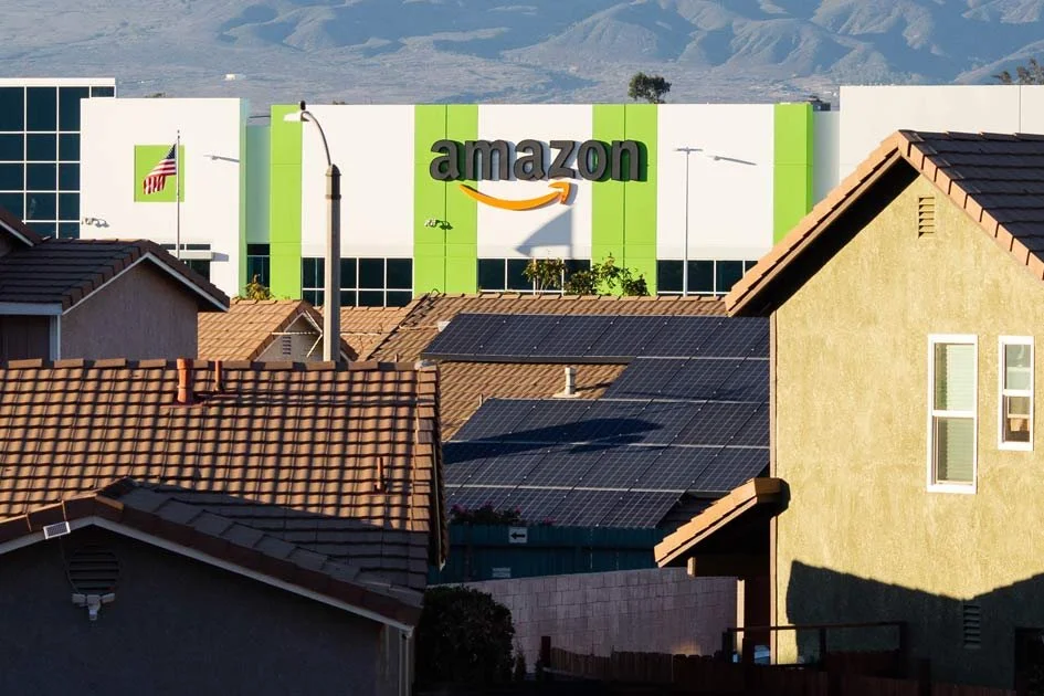 Residential neighborhood with houses and rooftops, some with solar panels, and a large Amazon warehouse in the background.