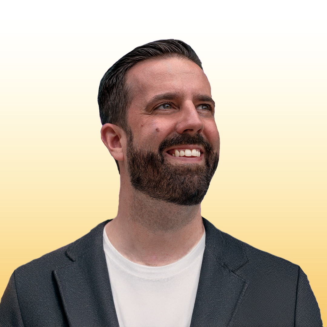 A smiling man with dark hair and a beard, wearing a dark blazer and a white t-shirt, looking slightly to the right with a neutral gradient background.