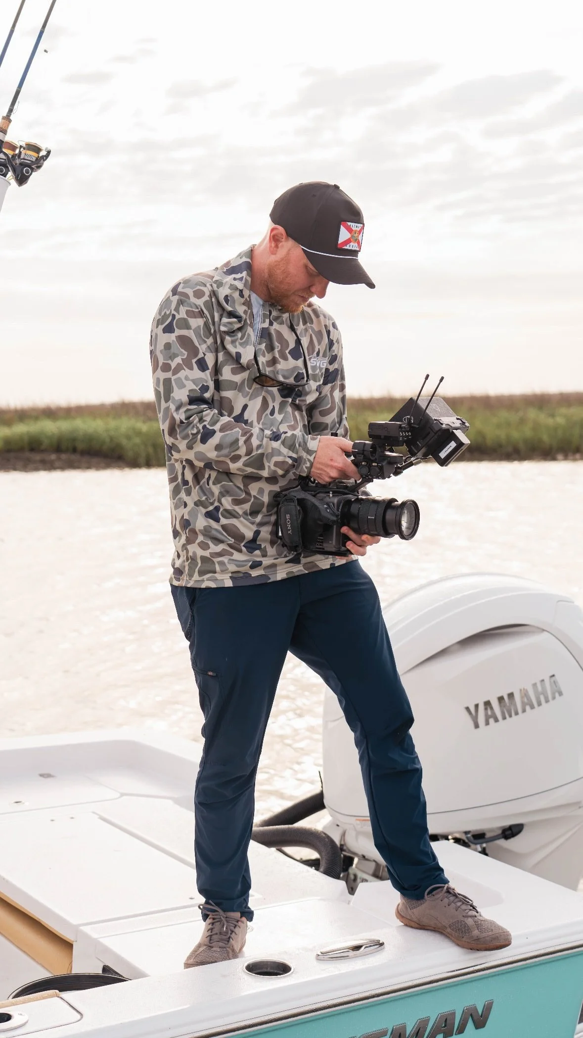A man standing on a boat holding a camera and filming. He wears a camouflage jacket, a black cap, and navy pants, with a Yamaha outboard motor behind him and an open water body in the background.