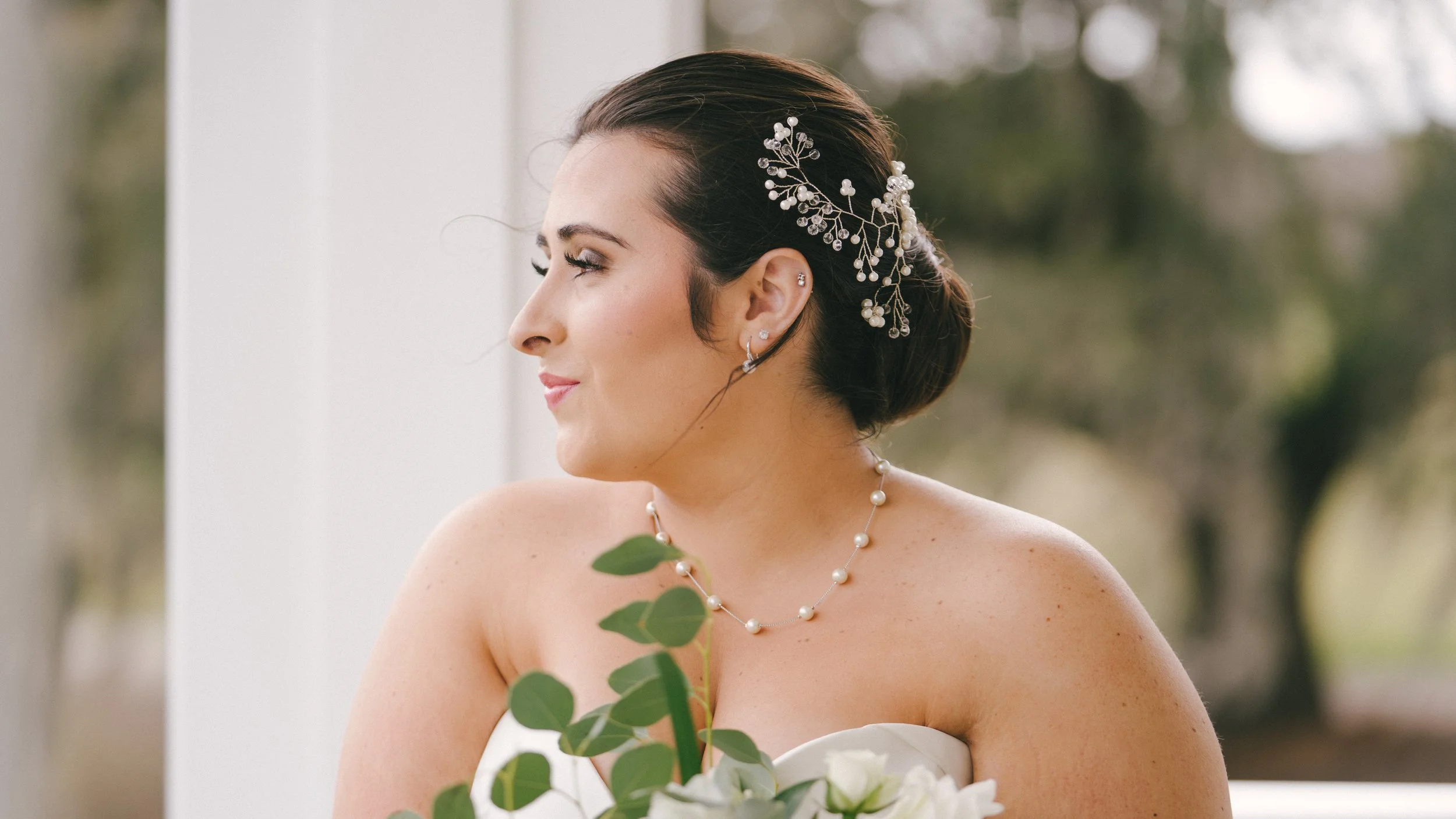 A bride with dark hair styled in an elegant bun, wearing a pearl and silver jewelry, holding a bouquet of white flowers and greenery, looking to her left with a joyful expression.