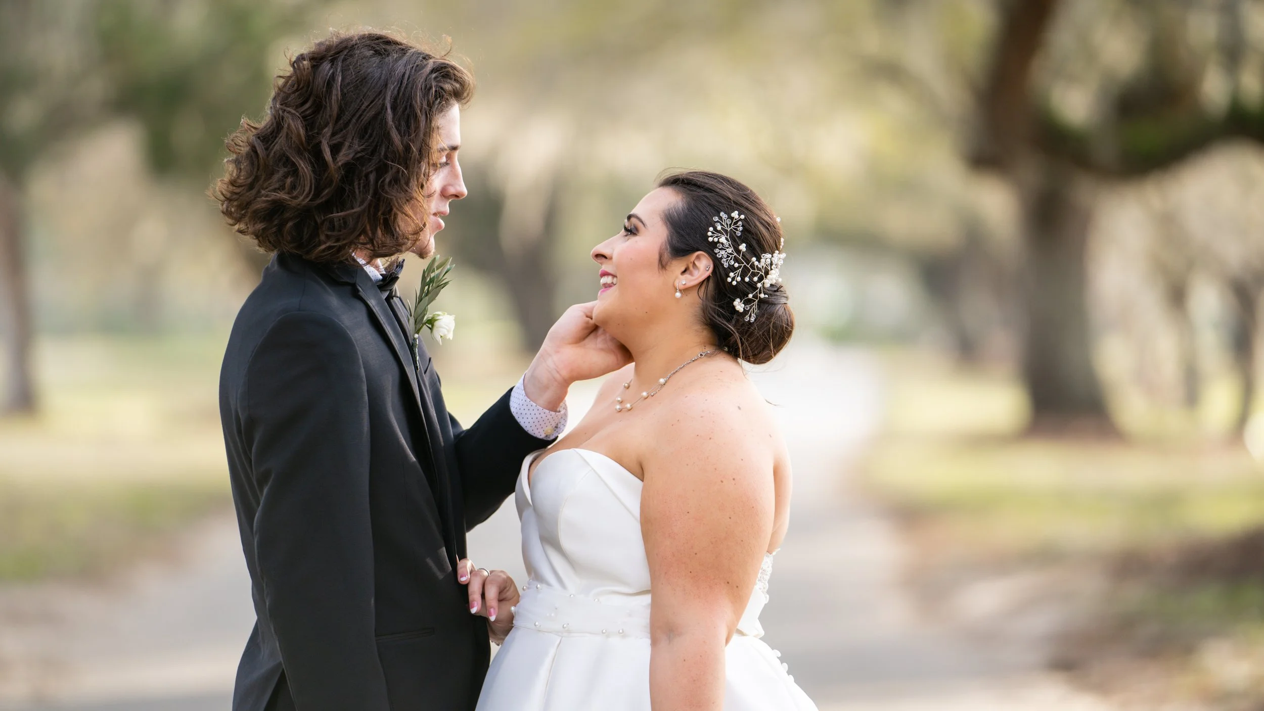 A bride and groom in wedding attire looking into each other's eyes outdoors in a park, with trees and a blurred pathway in the background.