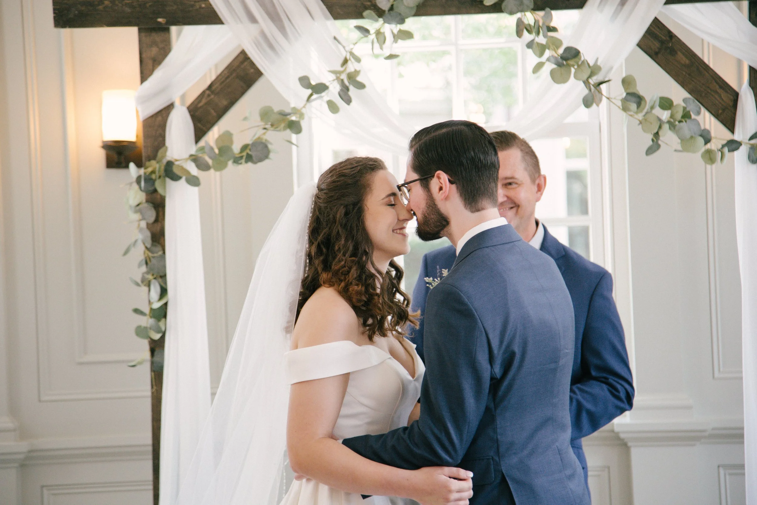 A bride and groom share a close, smiling moment at their wedding ceremony, standing beneath a wooden arch decorated with white drapery and eucalyptus leaves, with a cheerful officiant behind them.