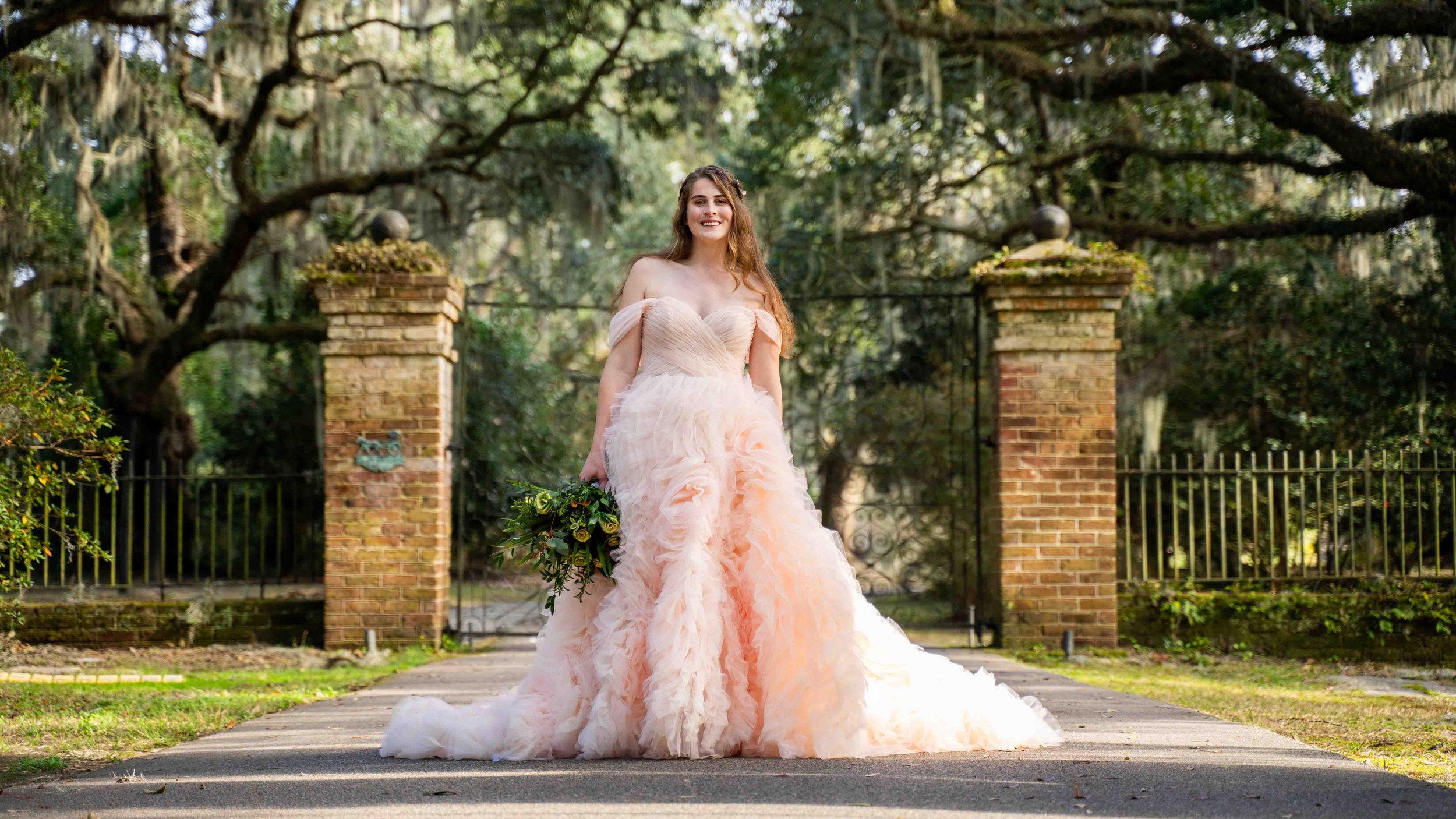A woman in a peach, ruffled, off-the-shoulder wedding gown holding a bouquet in front of a brick gate and large trees.