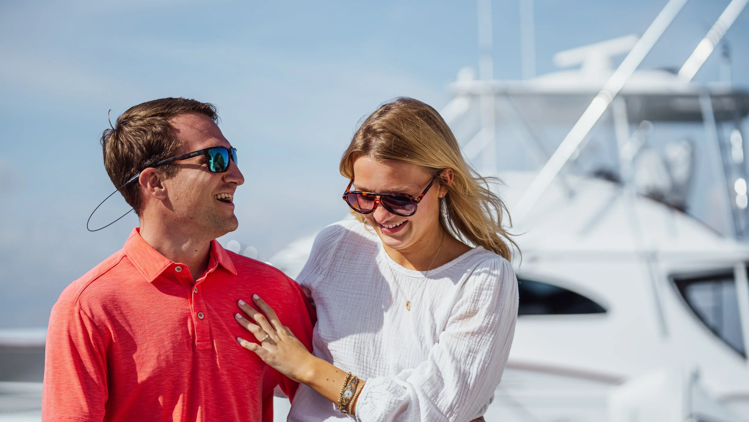 A man and woman smiling and laughing together near a white yacht, both wearing sunglasses on a sunny day.