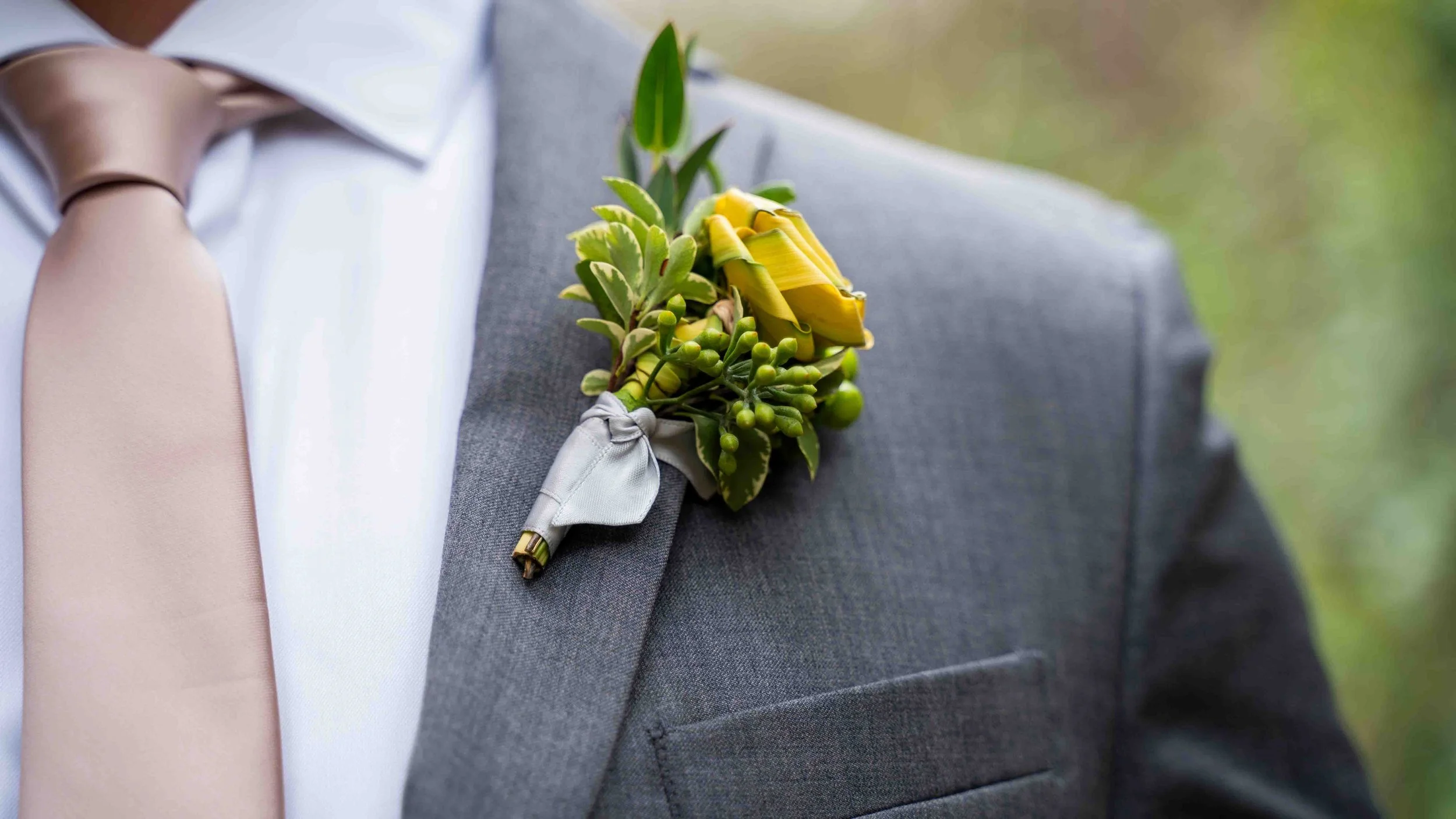 Close-up of a person's suit jacket with a yellow boutonniere, including a yellow rose and greenery, attached to the lapel.