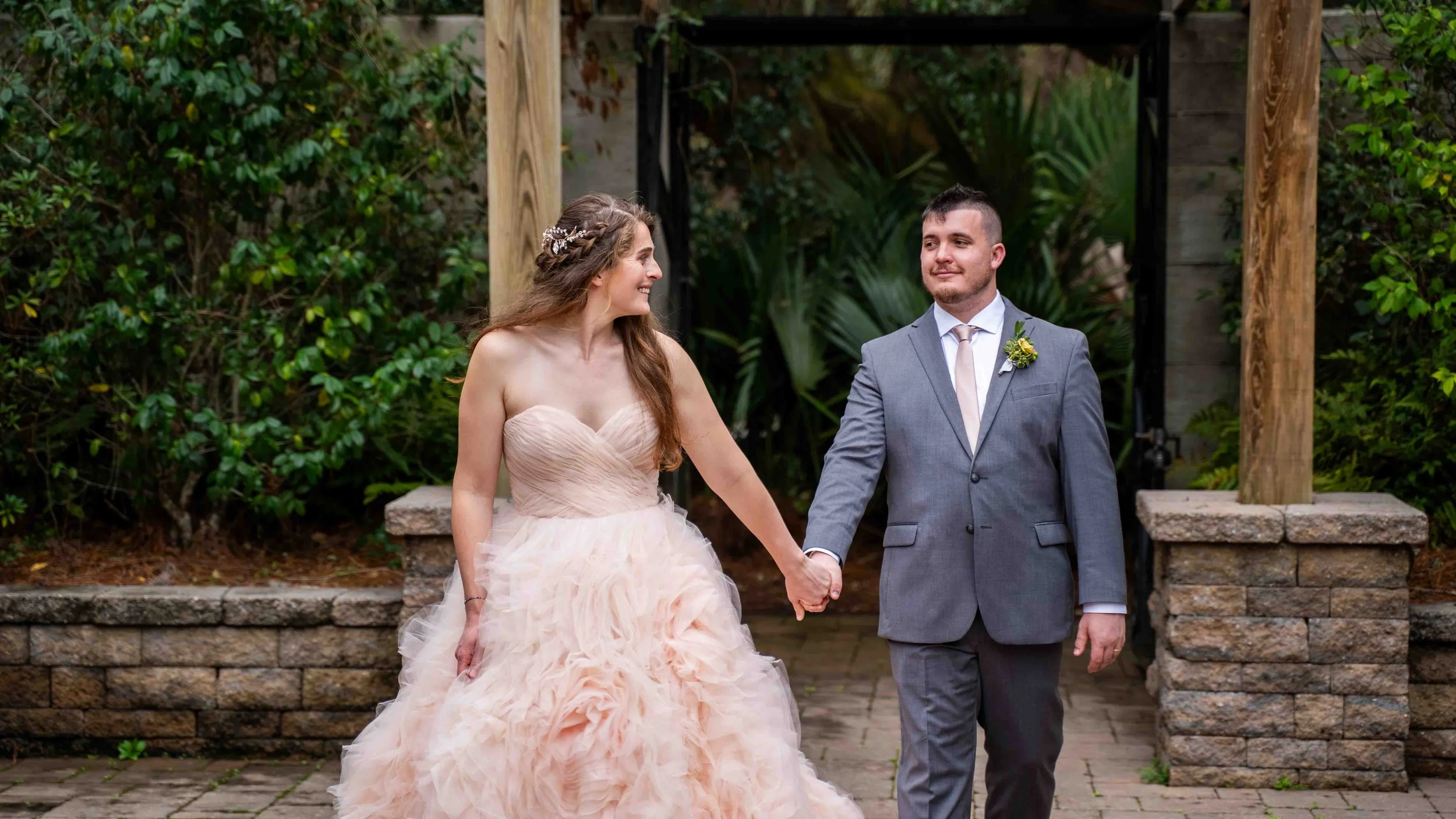 A bride and groom holding hands, walking outdoors on a wedding day. The bride is wearing a strapless, blush-colored wedding gown with a full, ruffled skirt, and has long, wavy hair adorned with a decorative hairpiece. The groom is dressed in a light 