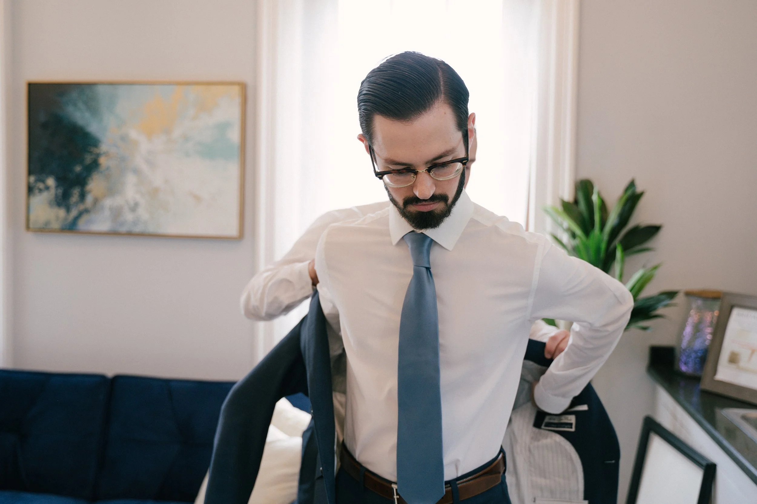 A man in a white shirt, blue tie, and glasses putting on a dark suit jacket in a bright room.