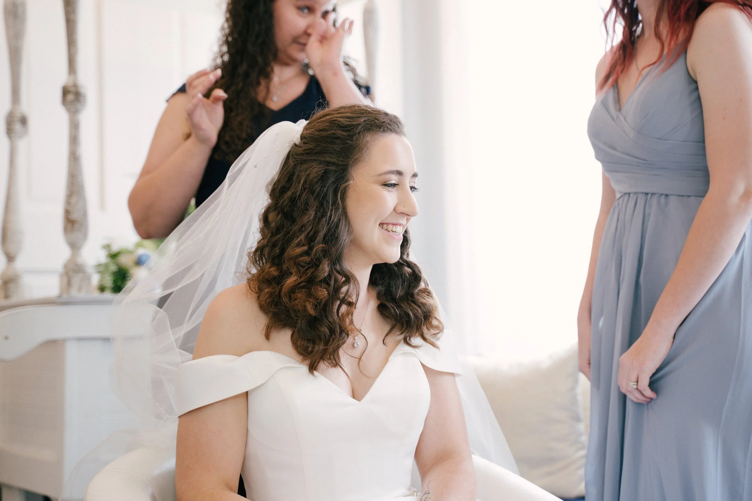 A bride with curly brown hair sitting in a white dress, smiling as her bridesmaids and a woman backstage prepare her for the wedding, including fixing her veil.