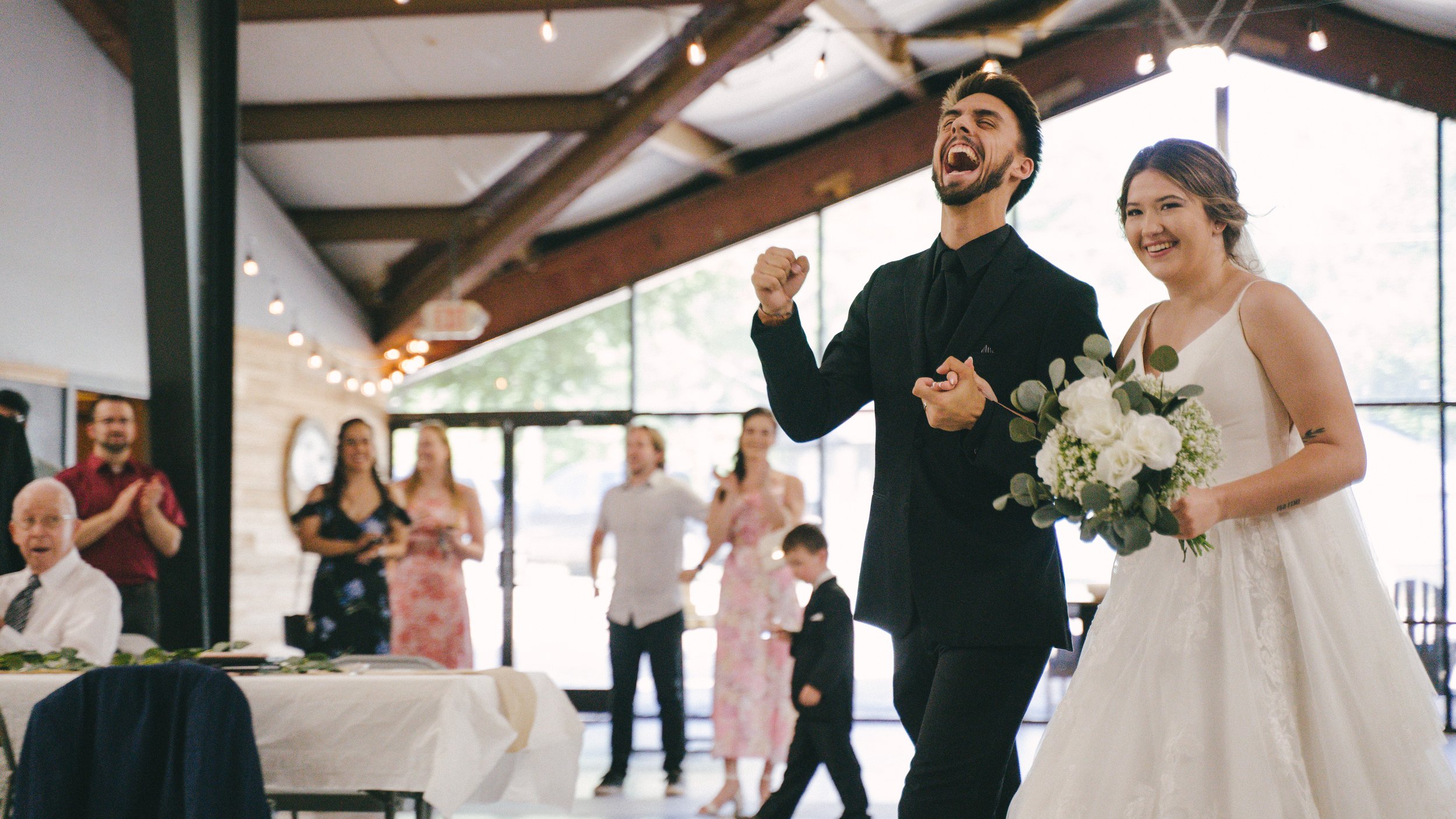 A bride and groom, both smiling broadly, holding hands and walking at their wedding reception. The bride is holding a bouquet of white flowers and green foliage, while the groom is dressed in a black suit. Guests in the background are clapping and wa