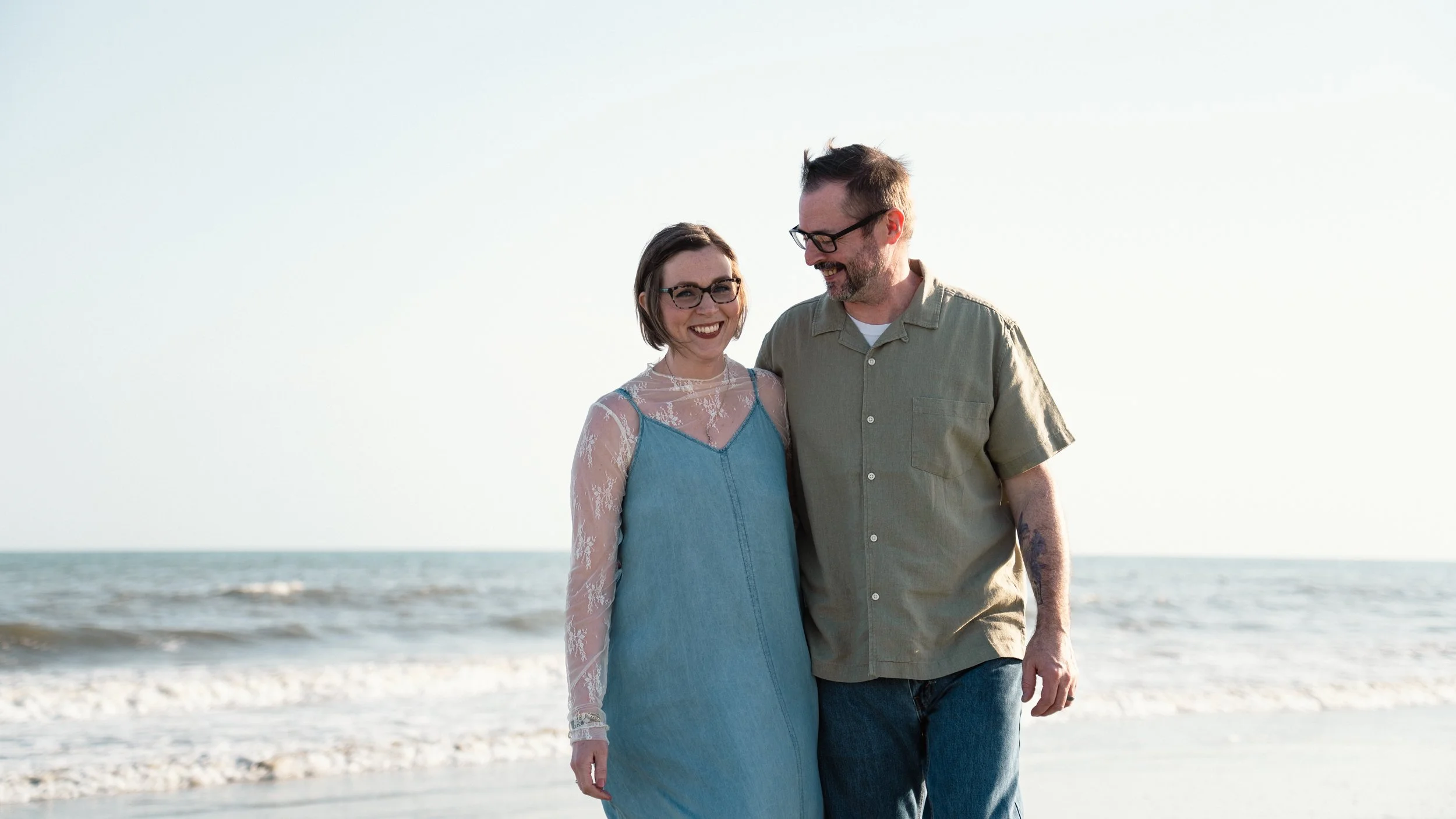 A smiling woman in a blue dress with lace sleeves and a man in a beige shirt walking on the beach together.