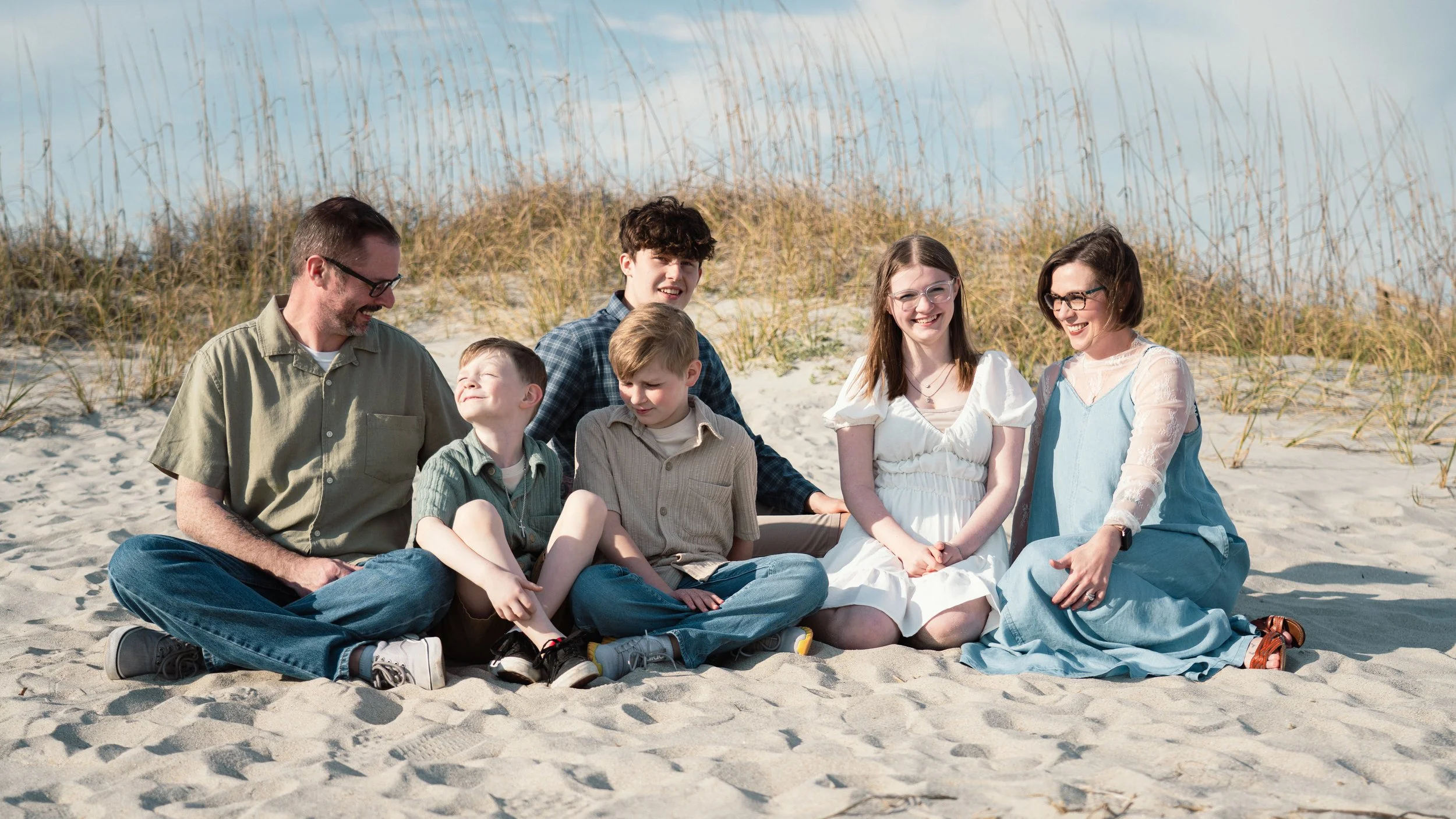 A family of seven sitting on the sandy beach near dunes with grass, smiling and enjoying sunny weather.