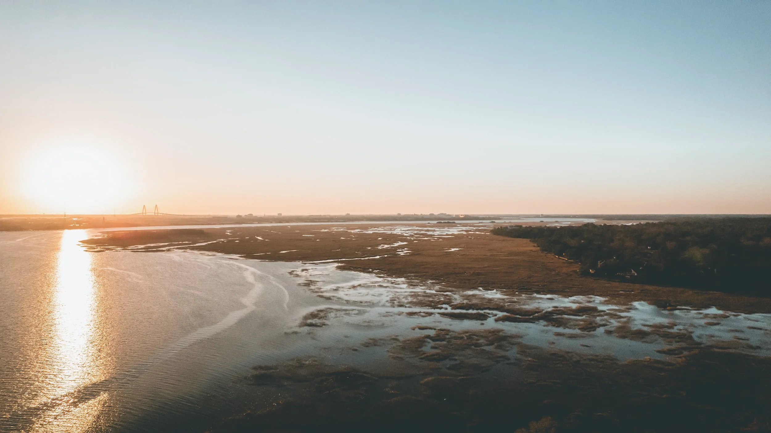 Sunset over a river with a bridge in the distance, dry land, and patches of greenery on the right.