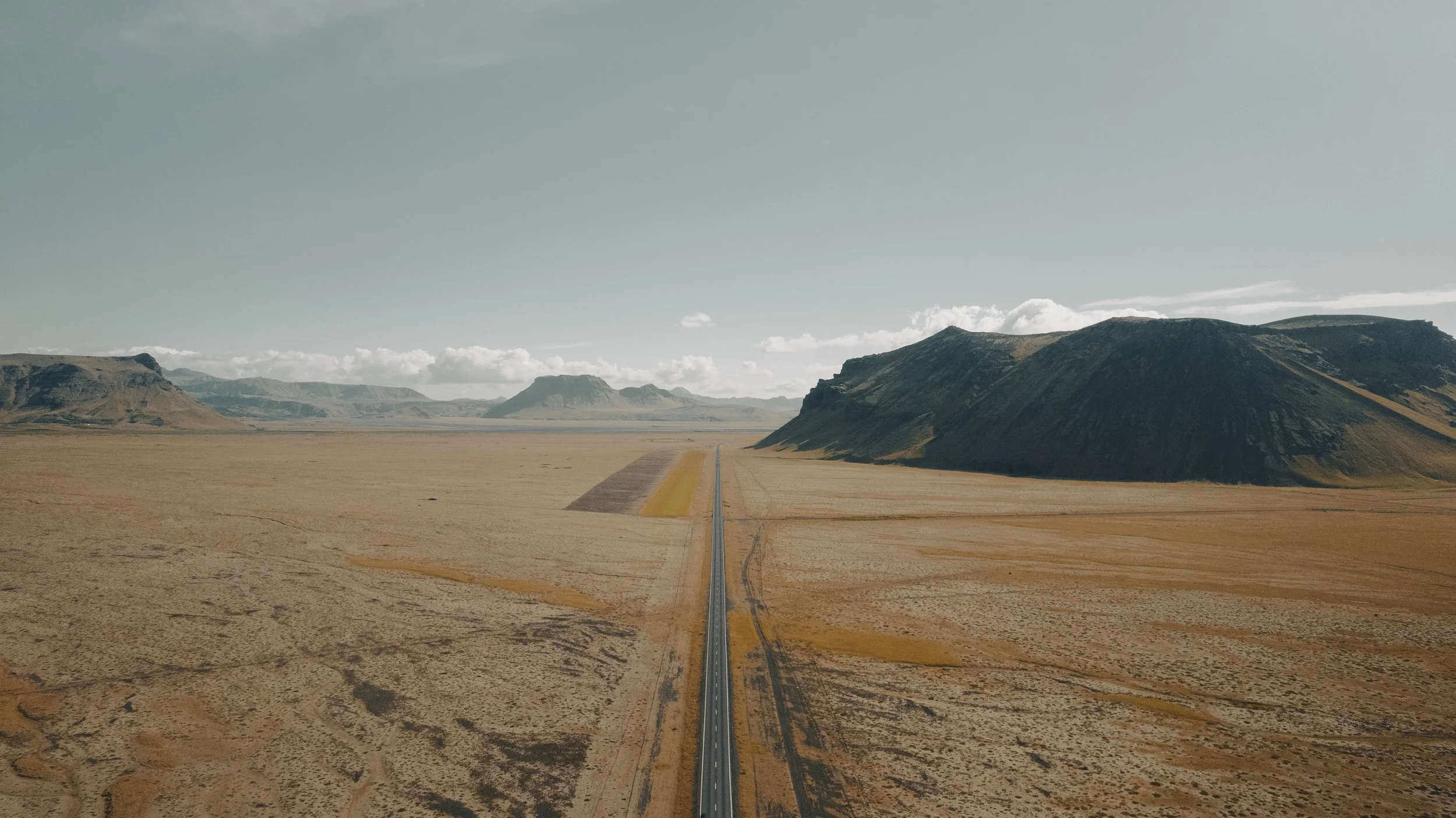 An expansive desert landscape with a long, straight road running through it, flanked by distant mountains under a partly cloudy sky.