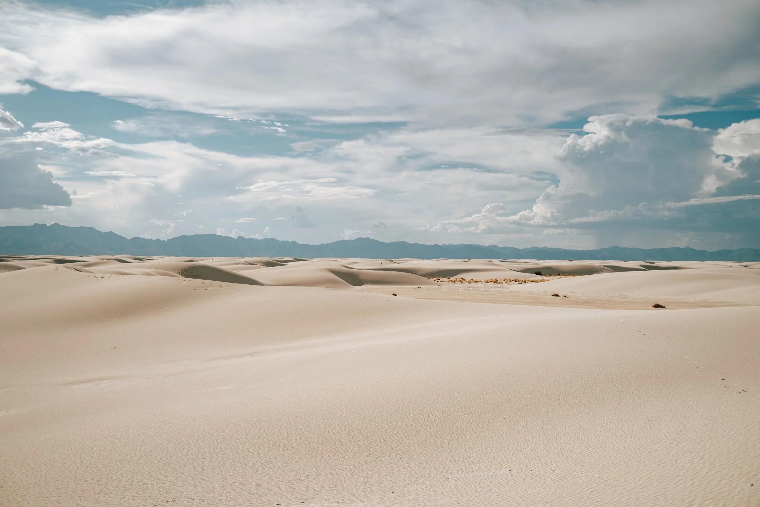 Expansive view of sand dunes under a partly cloudy sky, with mountain range in the distance.
