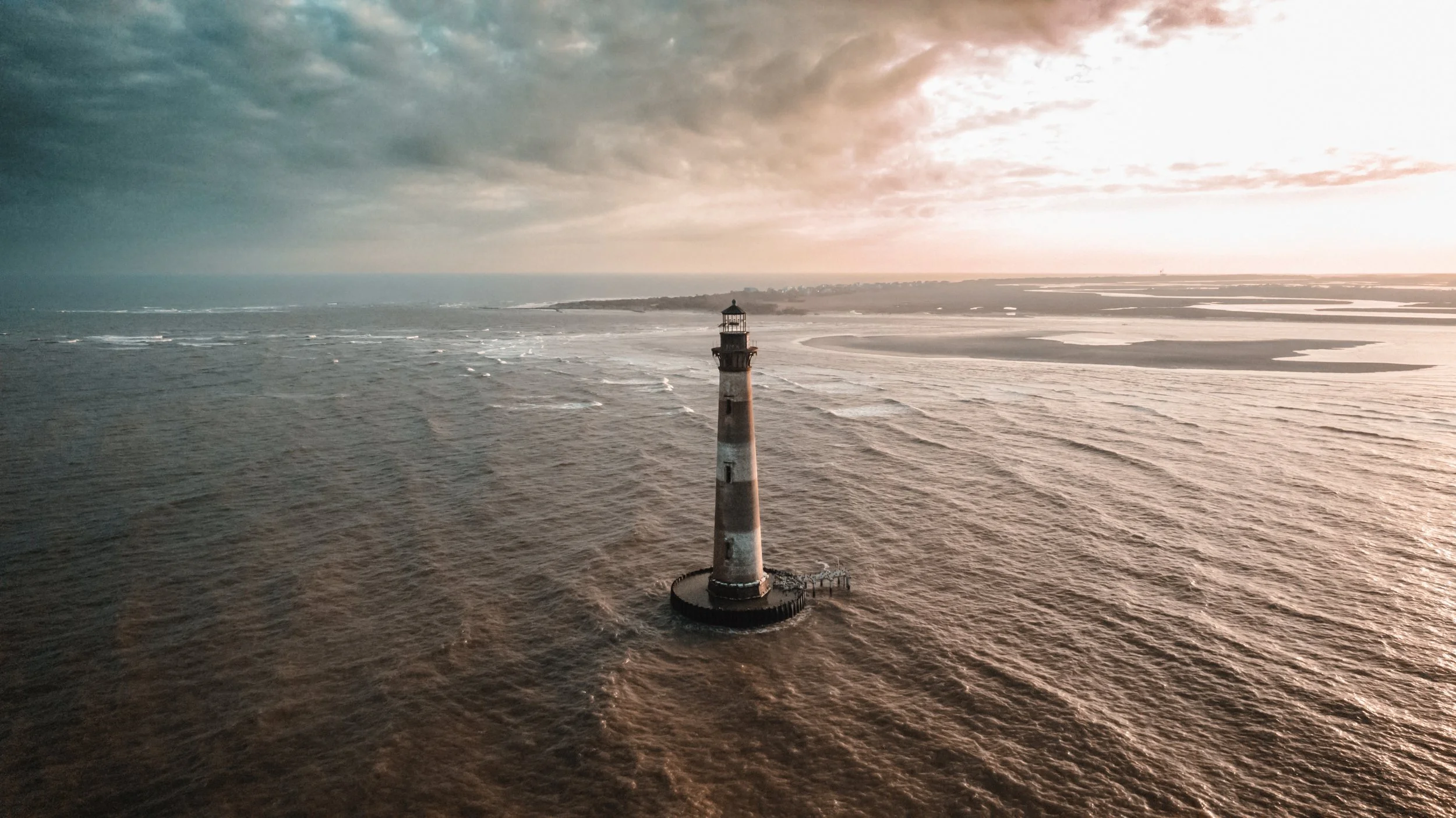 An aerial view of a lighthouse in the ocean during sunset or sunrise with cloudy sky in the background.