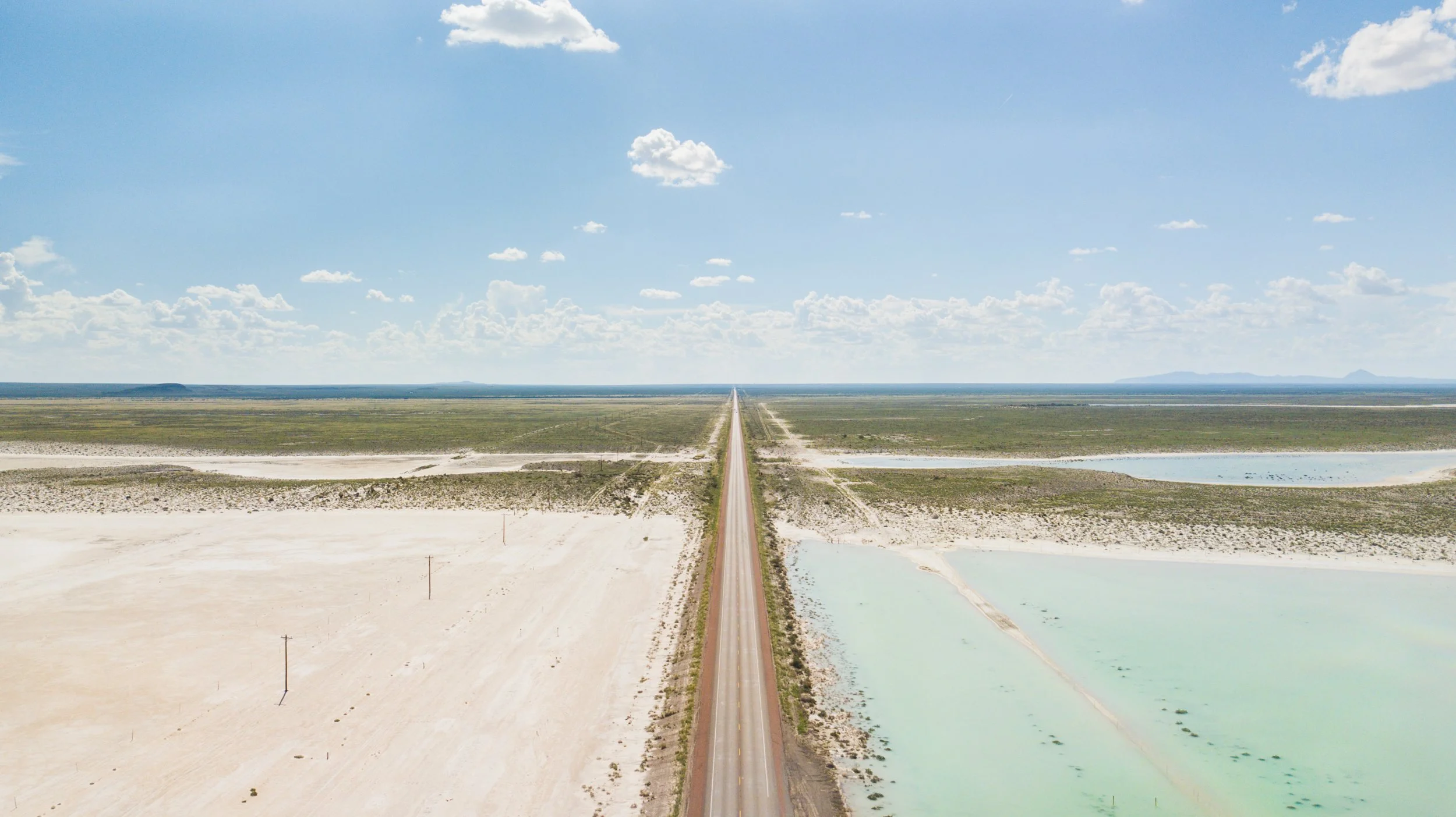 Aerial view of a long straight road running through a desert landscape with salt flats and water bodies on either side, under a partly cloudy sky.