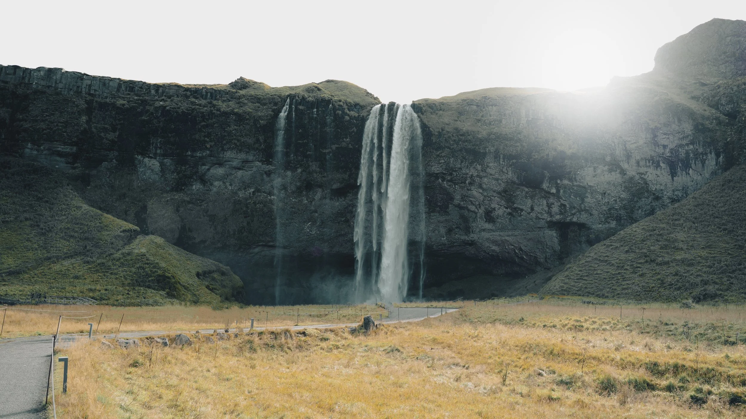 A large waterfall cascading down a high cliff into a valley with grassy fields, with sunlight coming from behind the cliff.