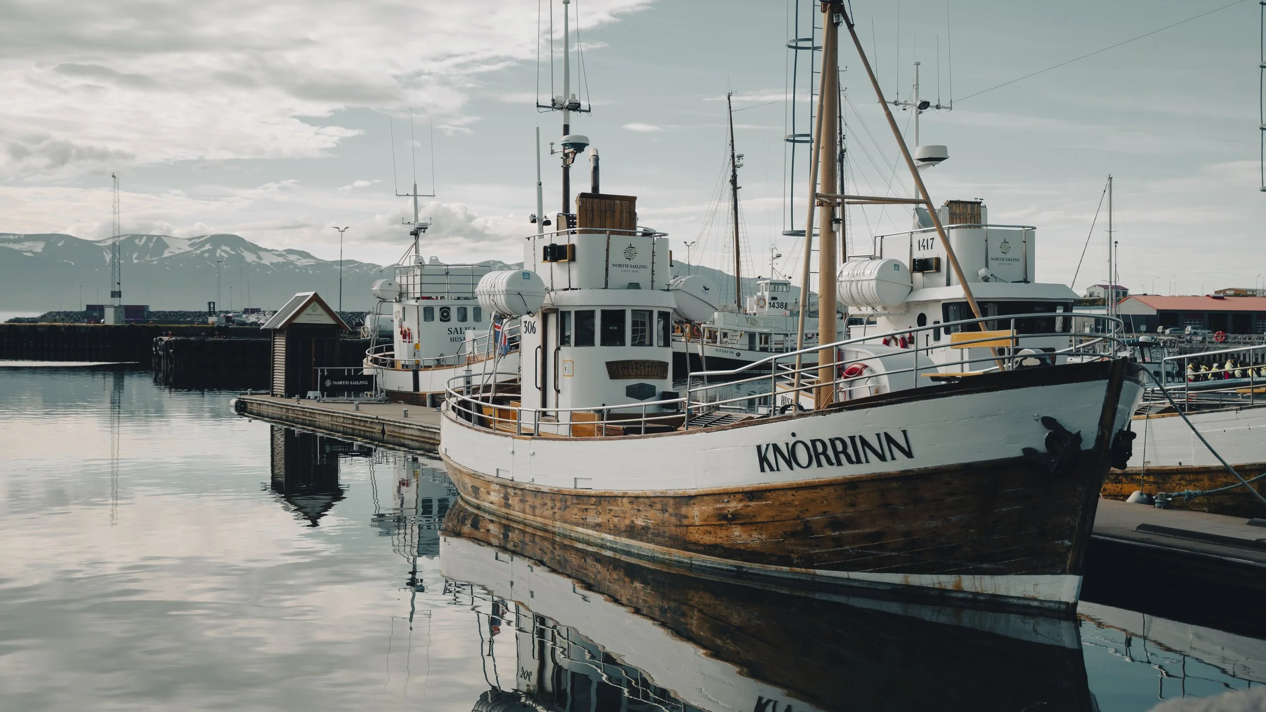 A docked boat named 'KNORRINN' at a marina with other boats, calm water, overcast sky, and mountains in the background.