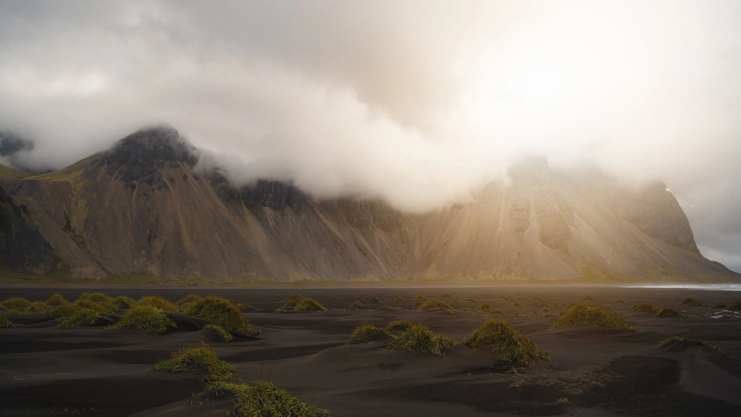 A black sand beach with scattered green mossy mounds in the foreground, tall mountain cliffs partially covered in low-hanging clouds in the background, soft sunlight illuminating part of the landscape.