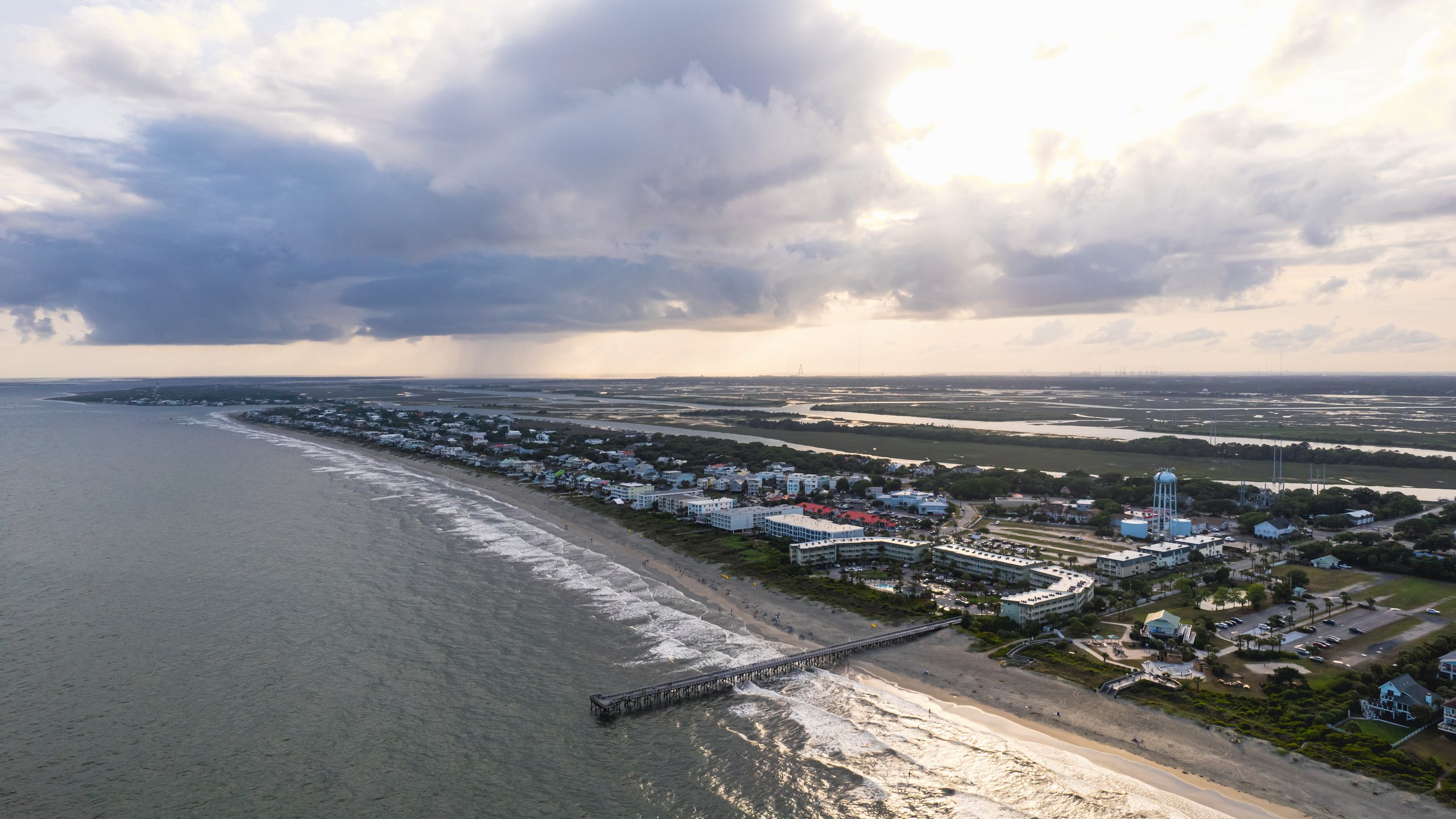 Aerial view of a coastal city with a beach, pier, residential buildings, and a water tower under cloudy sky.