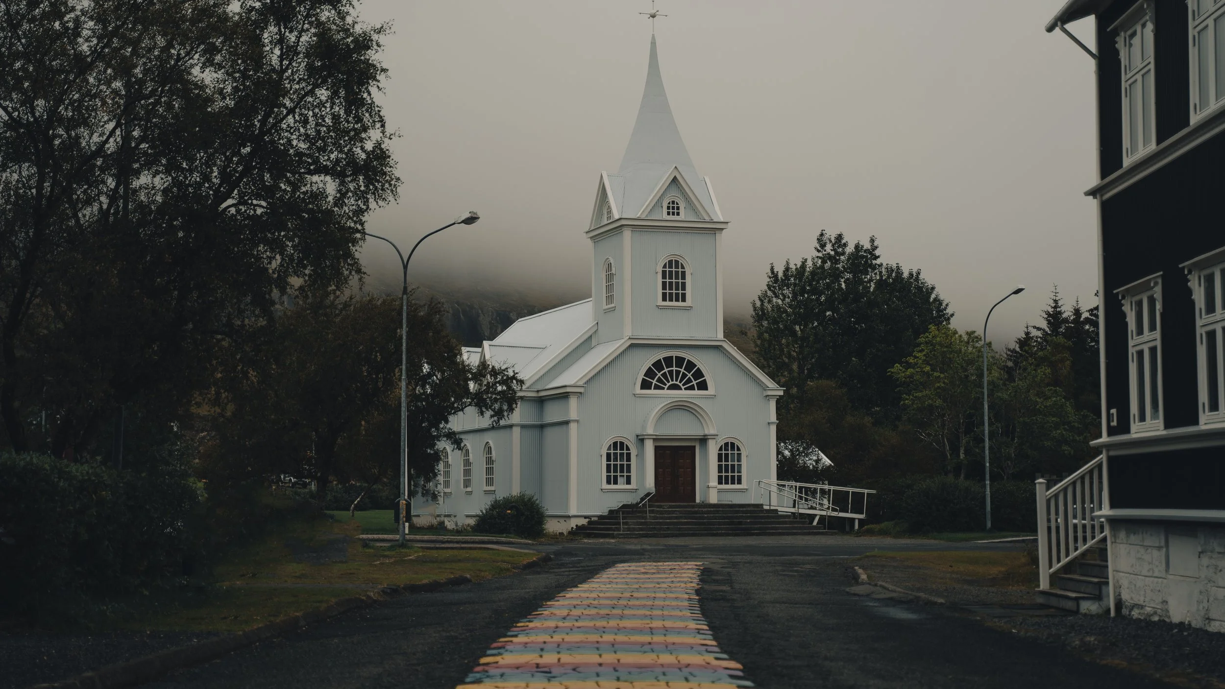 A white church with a tall steeple, steps leading up to the entrance, surrounded by trees and neighboring buildings, under a cloudy, foggy sky.