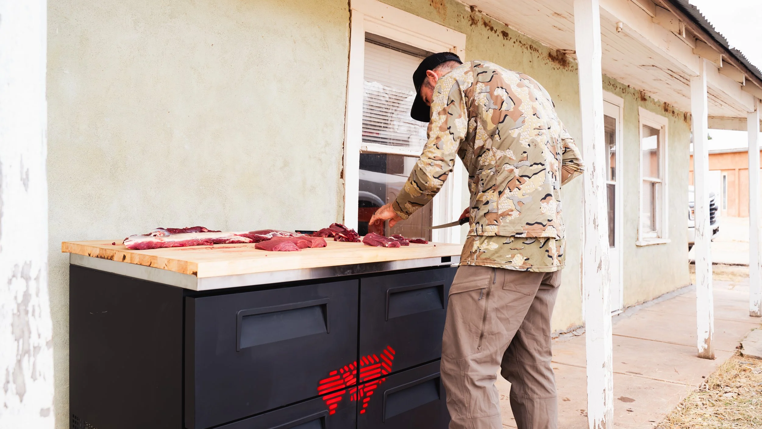 Man in camouflage jacket and khaki pants butchering meat on outdoor wooden table